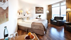 A hotel room with a bed, bedside lamps, a sofa, a dining table with fruit, and a painting on the wall. - Hotel Plöner See by Tulip Inn (Ploen)
