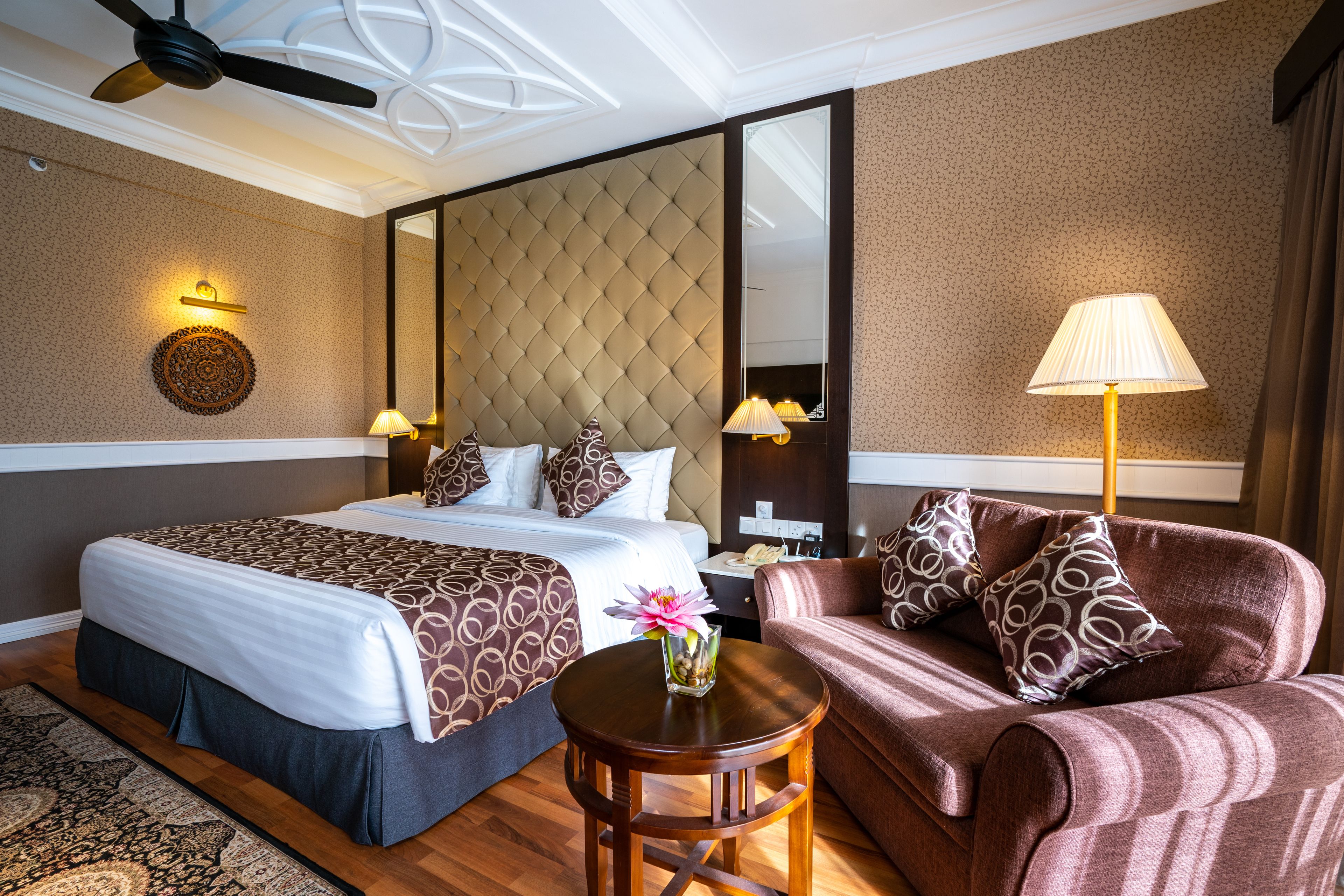 A hotel room with a large bed, a sofa, a small table, a lamp, and a ceiling fan.