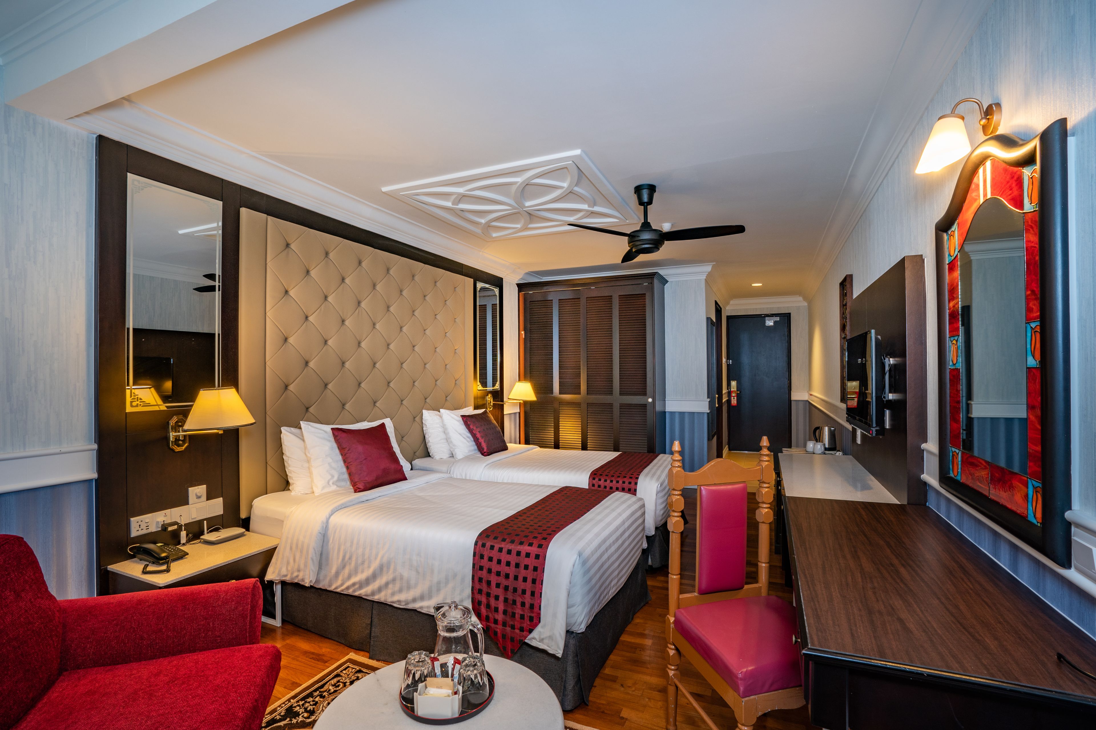 A hotel room with a large bed, a desk, a chair, a red armchair, and a ceiling fan.