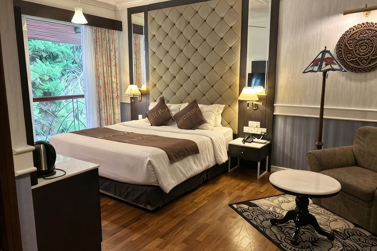 A hotel room with a large bed, a sitting area with a table, a coffee machine, and a view of greenery outside.
