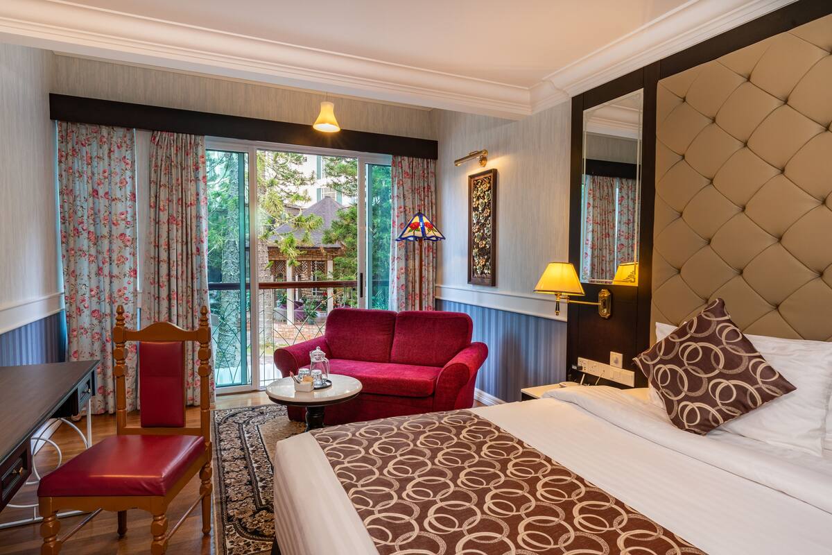Una habitación de hotel con cama, sofá rojo, silla de madera, mesita y ventana con cortinas.