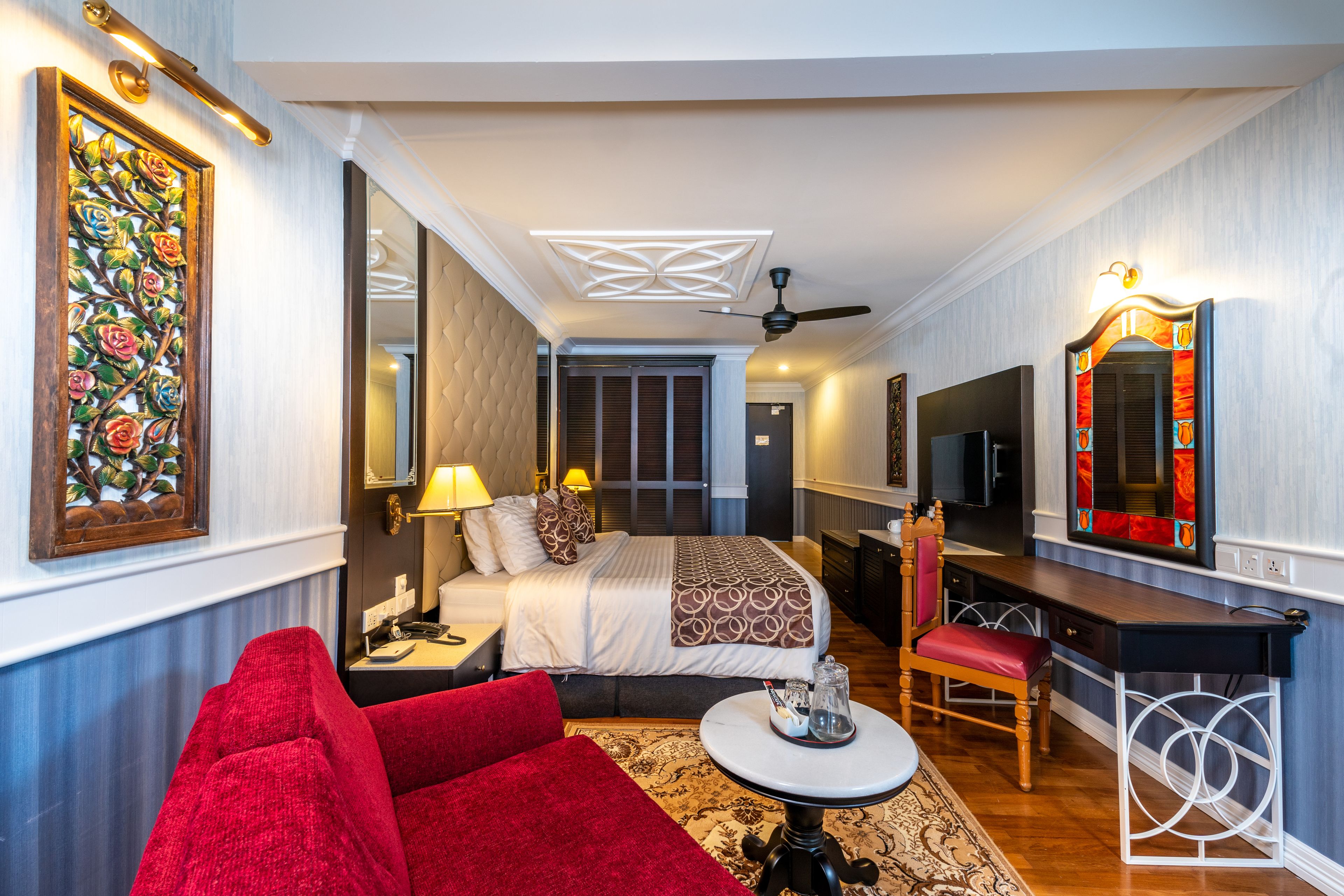A hotel room with a bed, a desk, a chair, a red sofa, a small table, and a mirror.