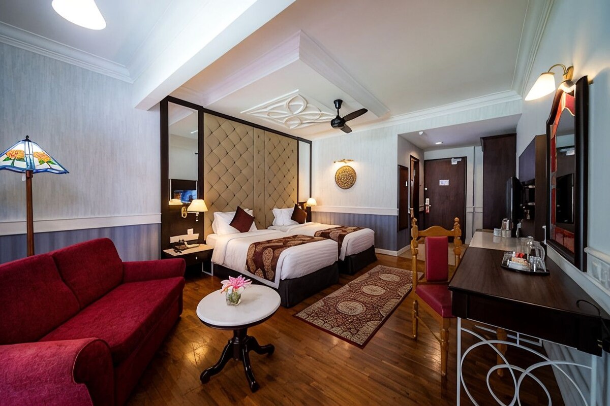 Une chambre d’hôtel avec un lit, un canapé rouge, une petite table avec un vase, un bureau avec un miroir et un ventilateur de plafond.