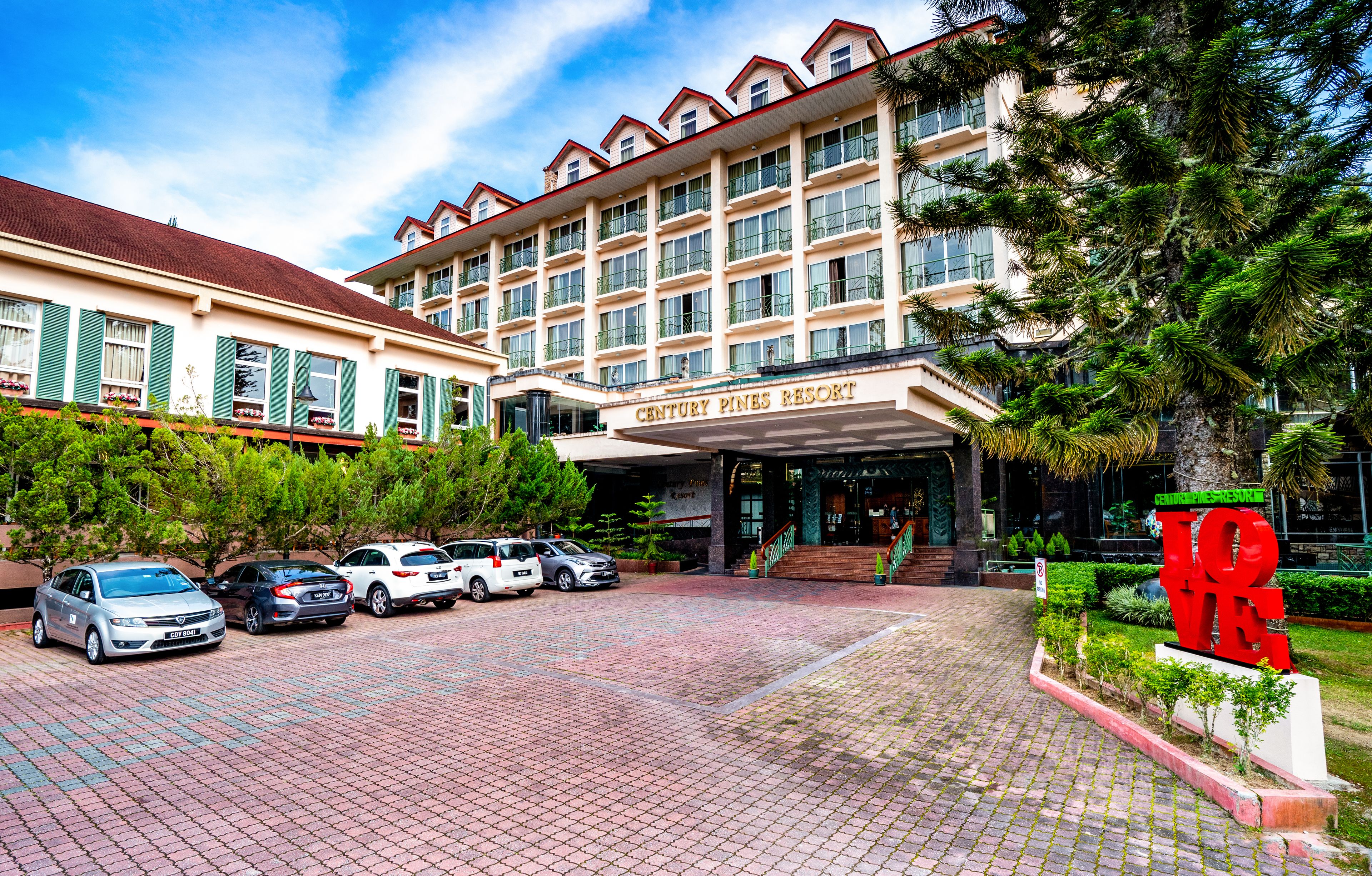 Un hotel con fachada roja y blanca, un acceso cubierto y un estacionamiento con varios automóviles.