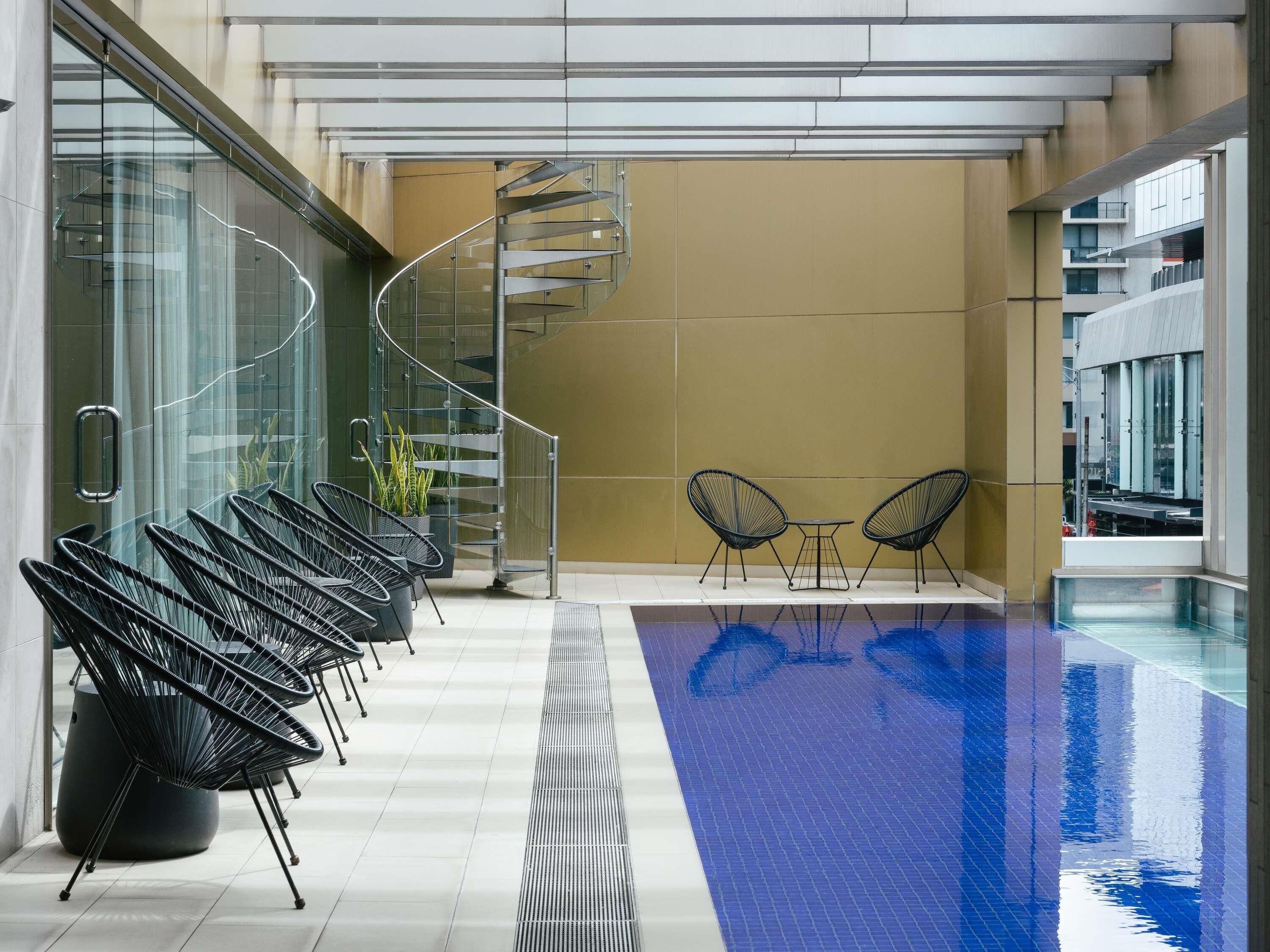 A modern indoor swimming pool with a glass railing, a spiral staircase, and black wireframe chairs.