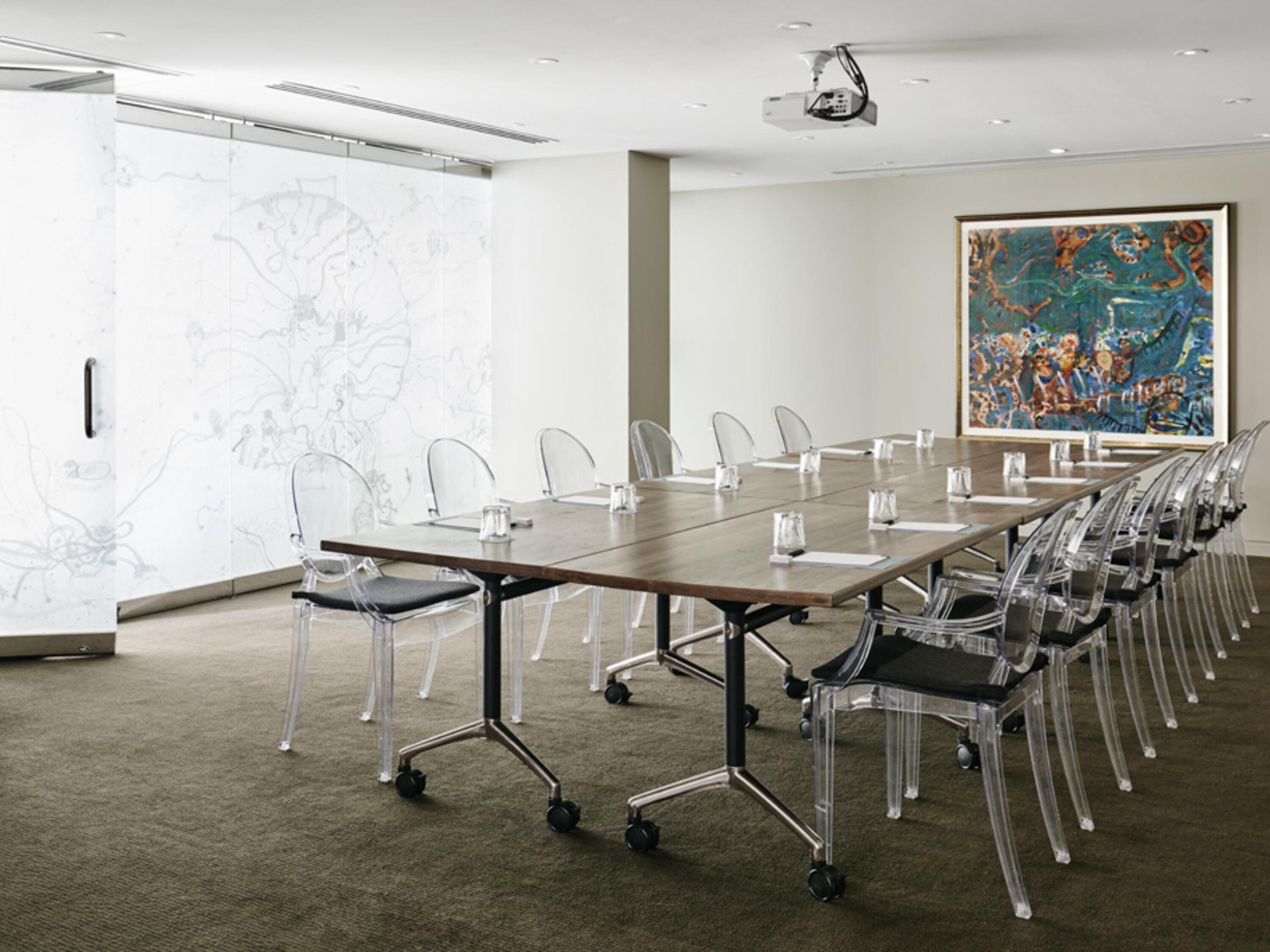 A conference room with a long table, clear chairs, a projector, and a large abstract painting on the wall.