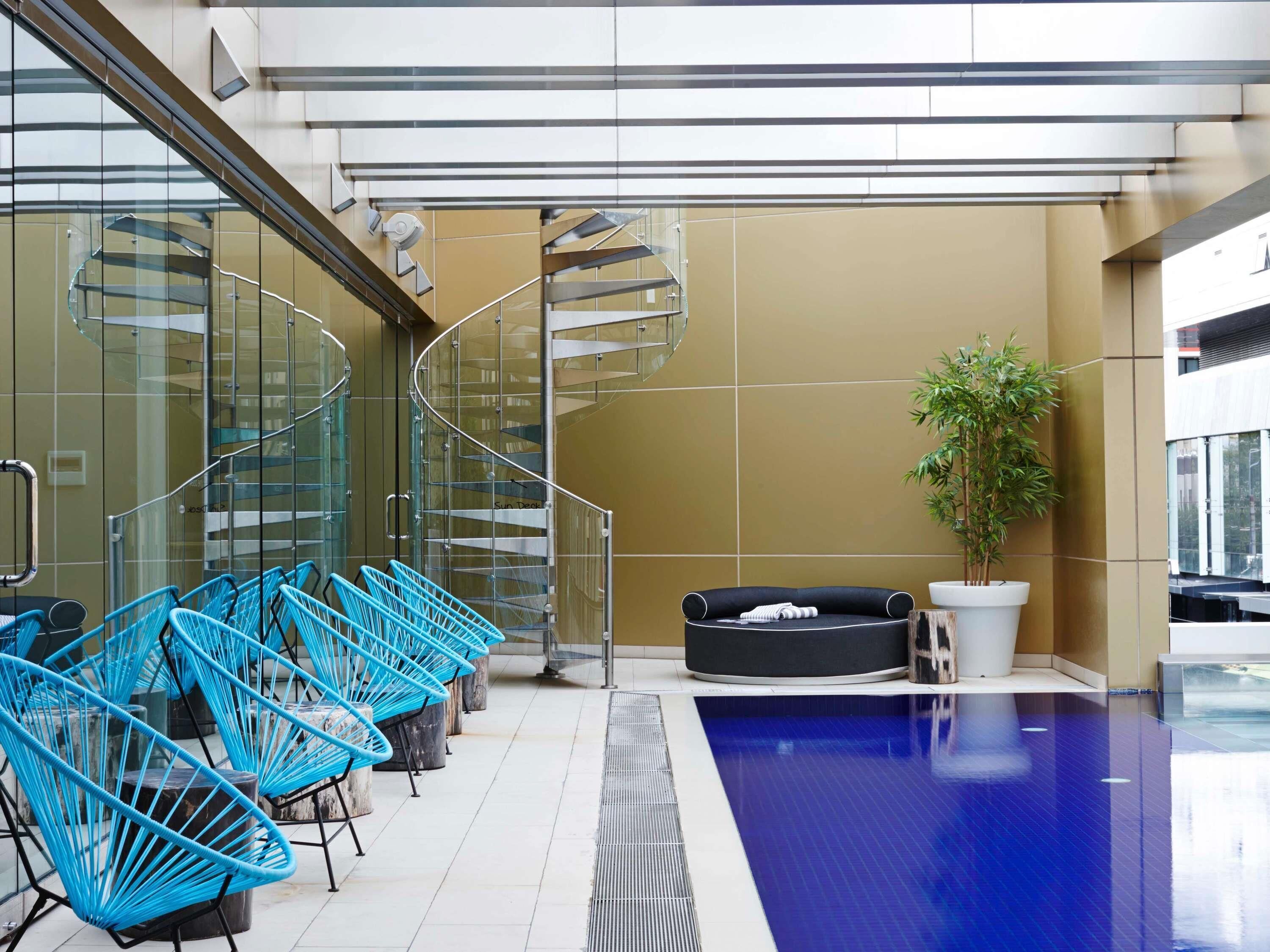Un área de piscina interior moderna con una escalera de caracol, sillas azules, un sofá negro y una planta en maceta.
