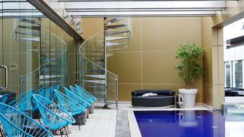 Un área de piscina interior moderna con una escalera de caracol, sillas azules, un sofá negro y una planta en maceta.