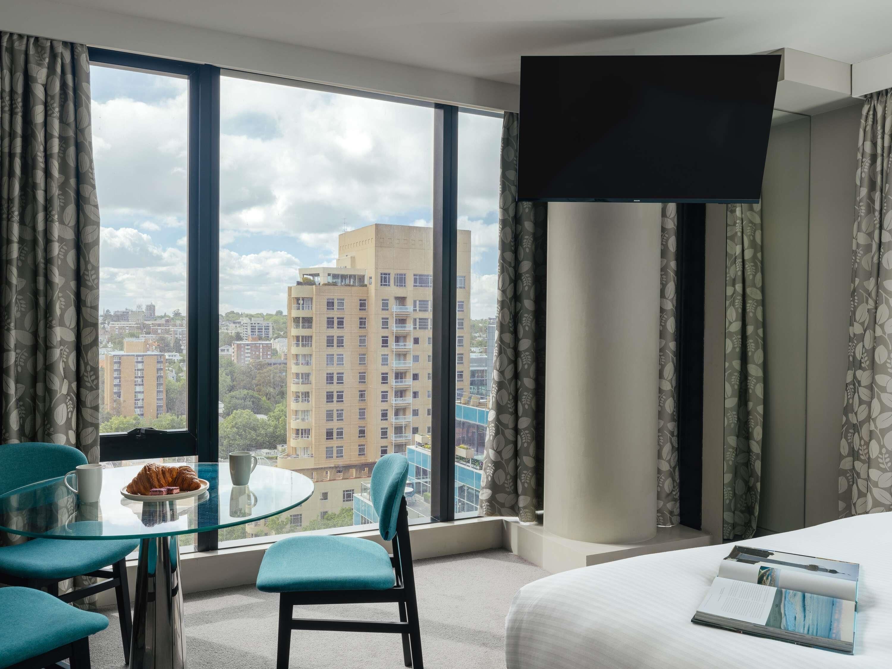 Une chambre d’hôtel avec une grande fenêtre, un lit, une table avec un croissant et une chaise.