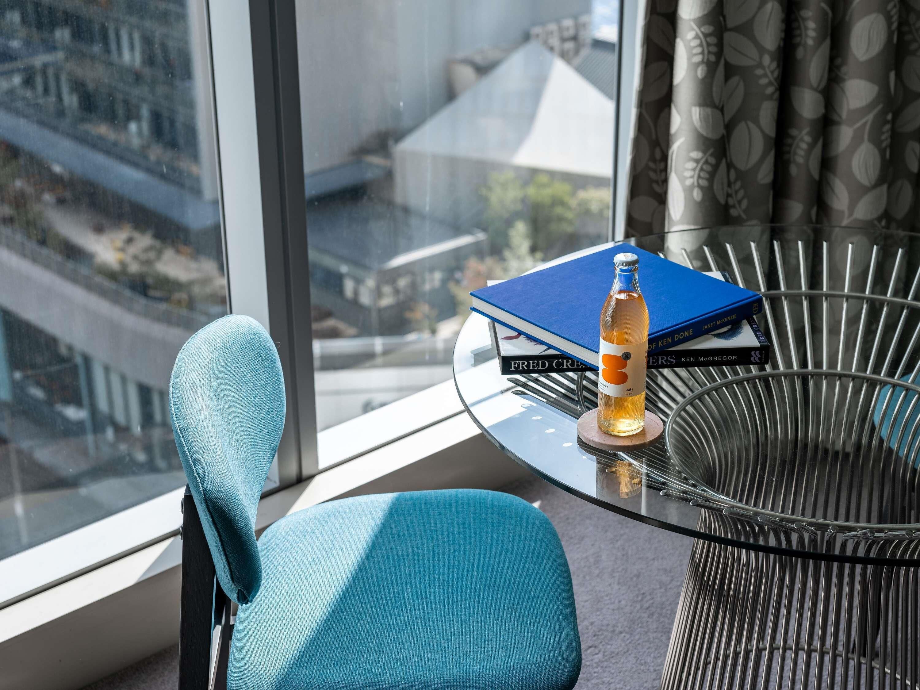 A room with a view of buildings, a blue chair, a glass table with a bottle and books.