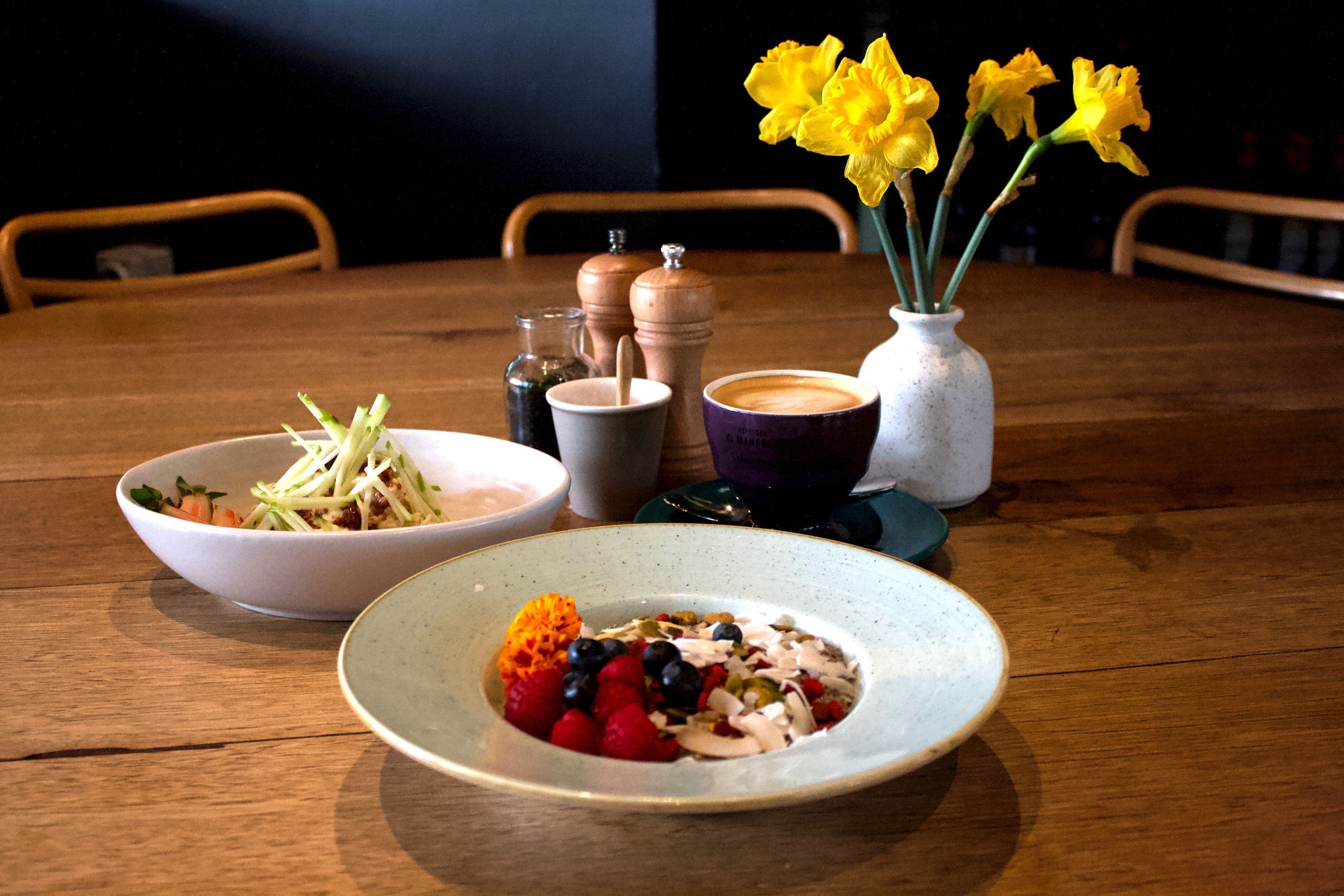 Una mesa de madera con una ensalada en un bol, un plato de fruta, una taza de café y un jarrón con flores amarillas.