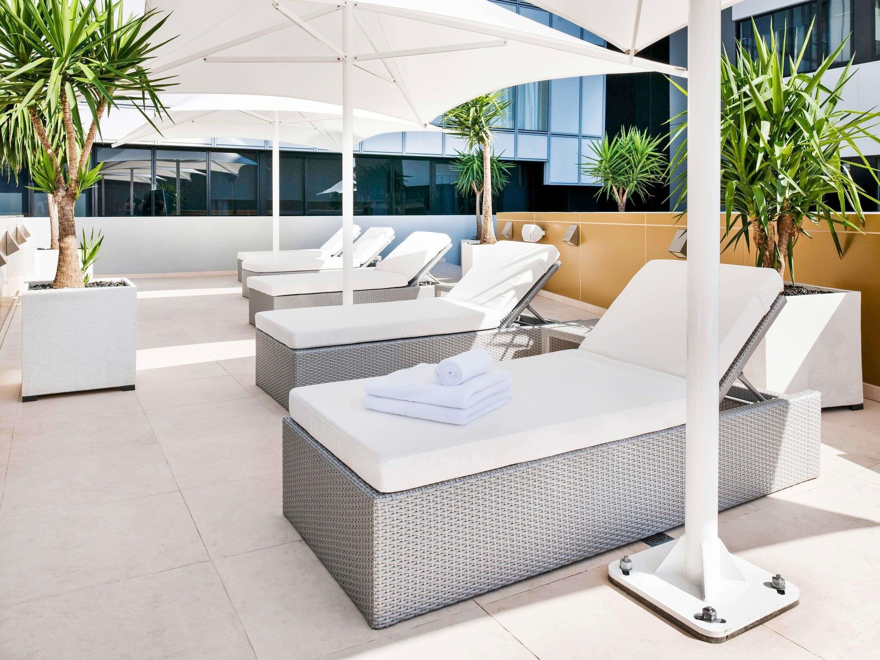 Uma área de lounge no terraço com espreguiçadeiras acolchoadas em branco, um guarda-sol branco e plantas em vasos.