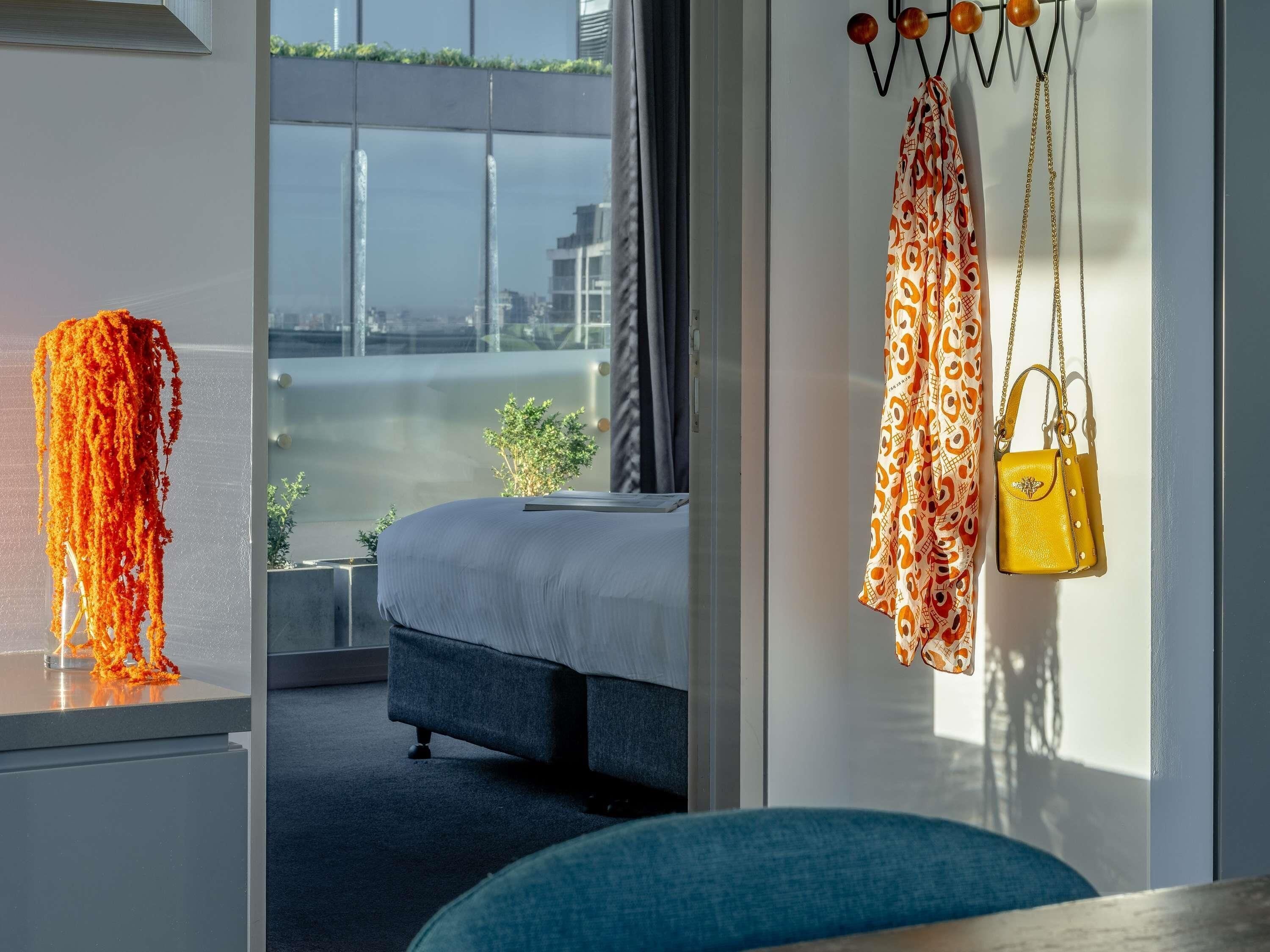 A modern hotel room with a bed, a window with a view of the city, and a hanging yellow handbag.