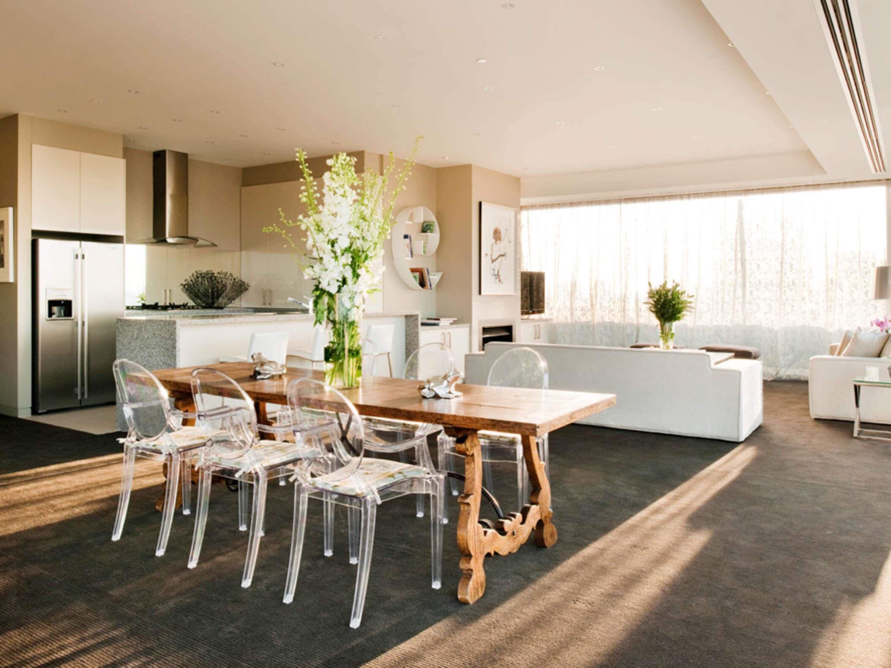 Una cocina moderna con horno empotrado, un comedor con mesa de madera y sillas transparentes, y una sala de estar con sofá y un ventanal grande.