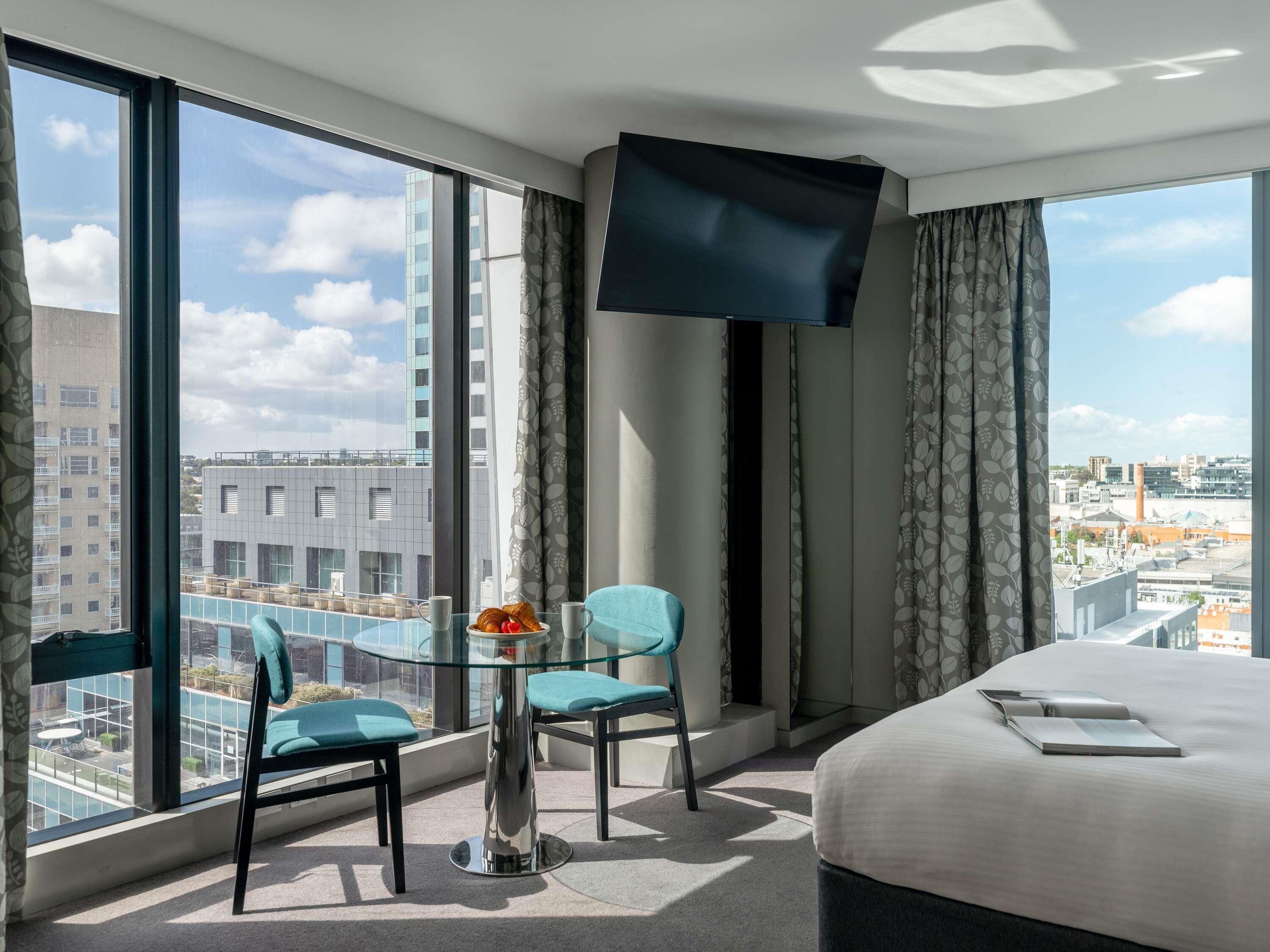 A hotel room with a bed, a TV, a small table with a glass top, two chairs, and a view of the cityscape through large windows.
