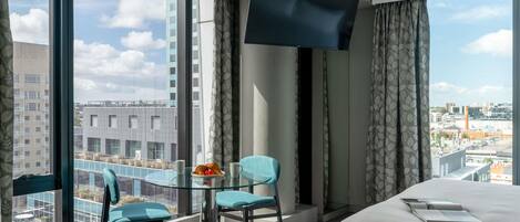 Habitación de hotel con cama, televisión, una mesita con superficie de vidrio, dos sillas y vistas a la ciudad a través de amplias ventanas.