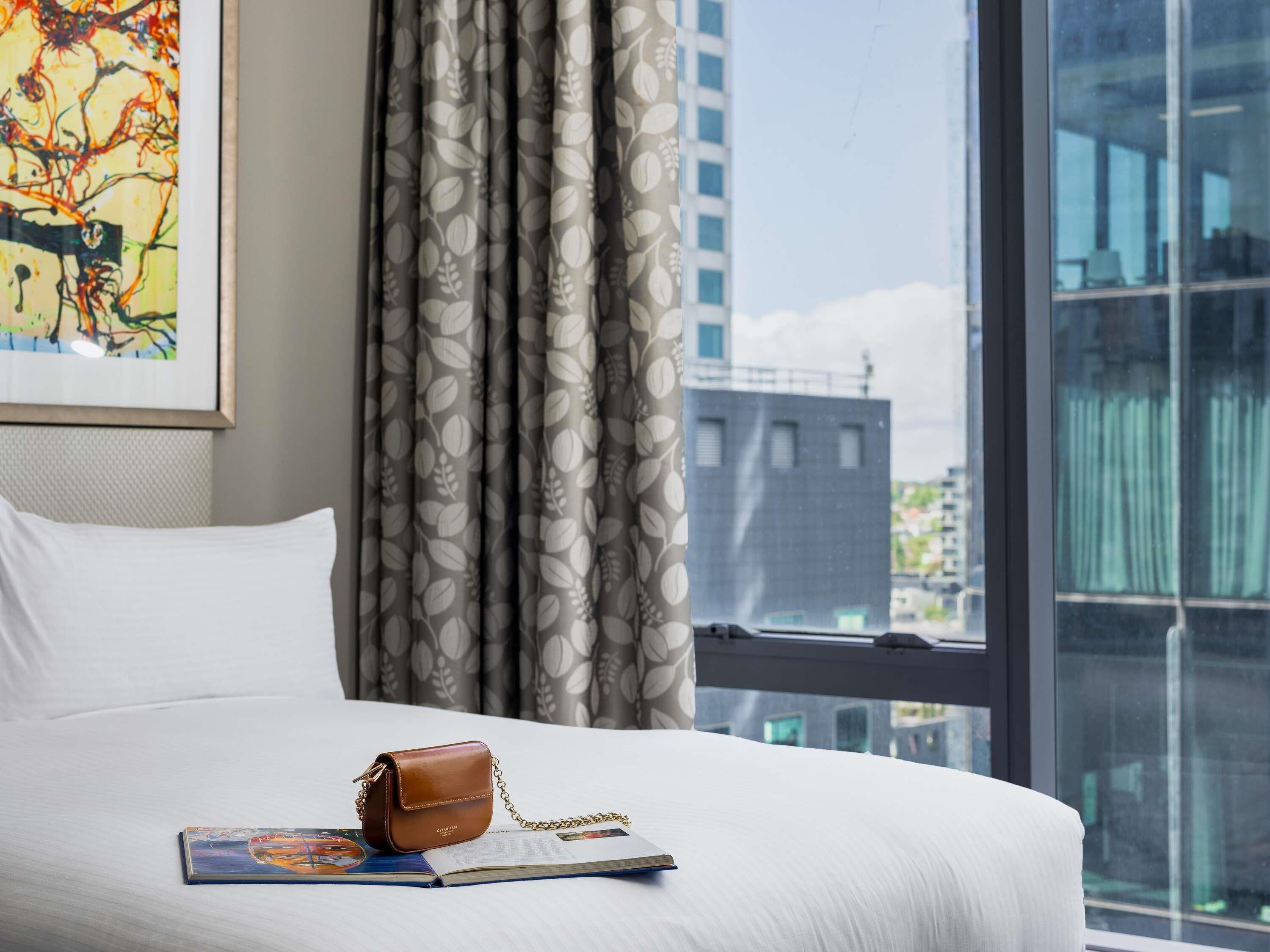 A hotel room with a bed, a window with a view of buildings, a curtain, and a brown handbag on the bed.