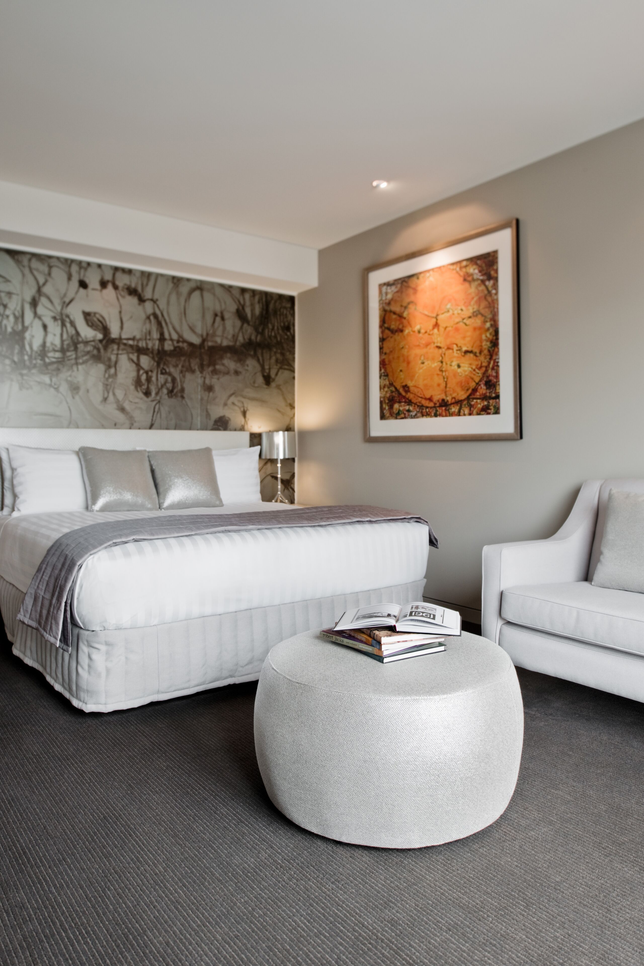 A modern bedroom with a large bed, a round ottoman, a white armchair, and a large abstract painting on the wall.