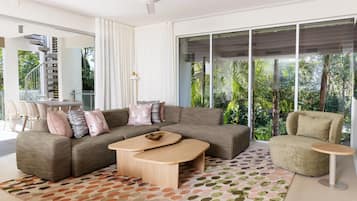 A modern living room with a sofa, coffee table, and armchair, large windows with a view of greenery, and a staircase.