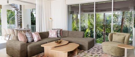 A modern living room with a sofa, coffee table, and armchair, large windows with a view of greenery, and a staircase.