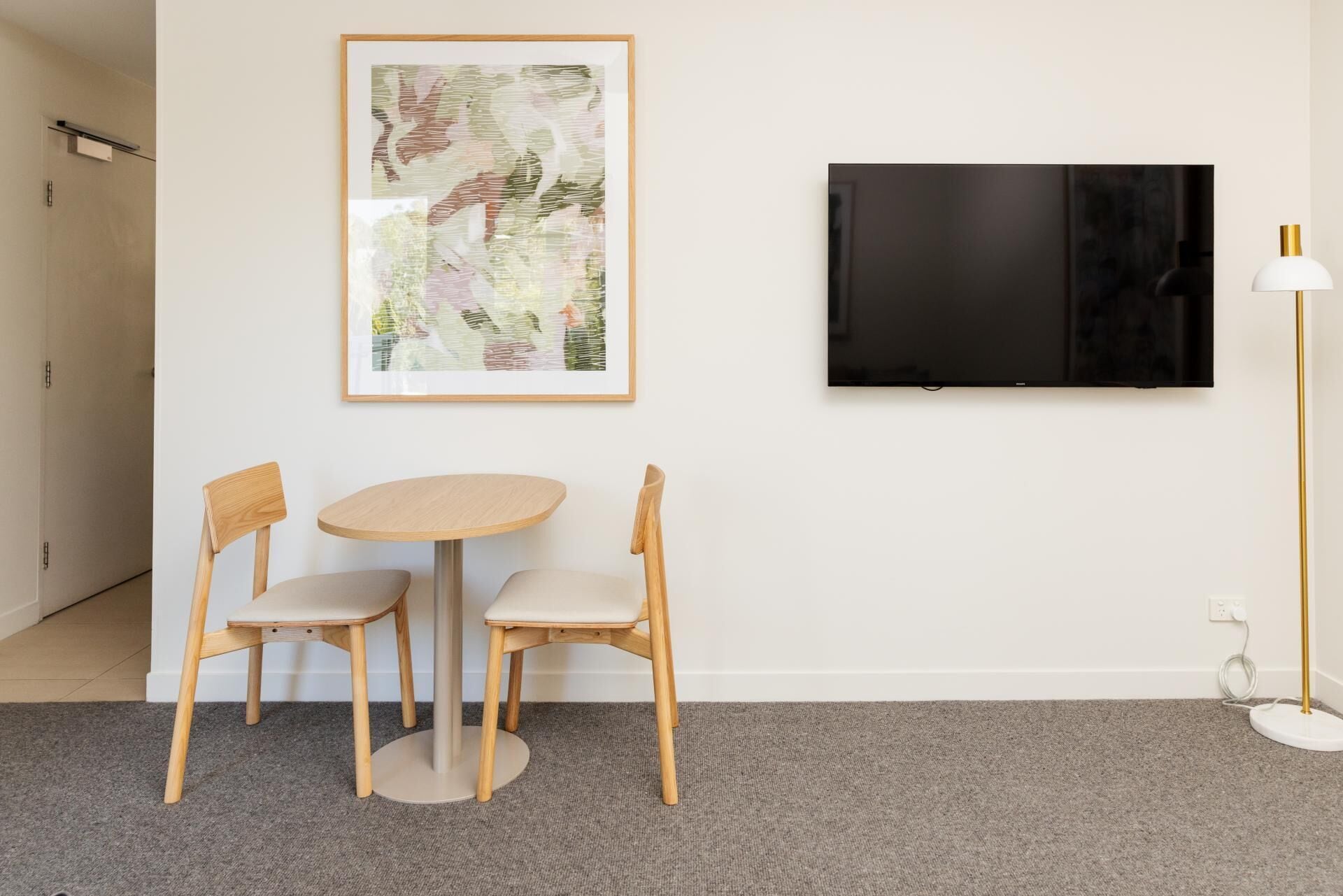 Una habitación con una mesa redonda, dos sillas de madera, un cuadro abstracto enmarcado, un televisor de pantalla plana y una lámpara de pie.