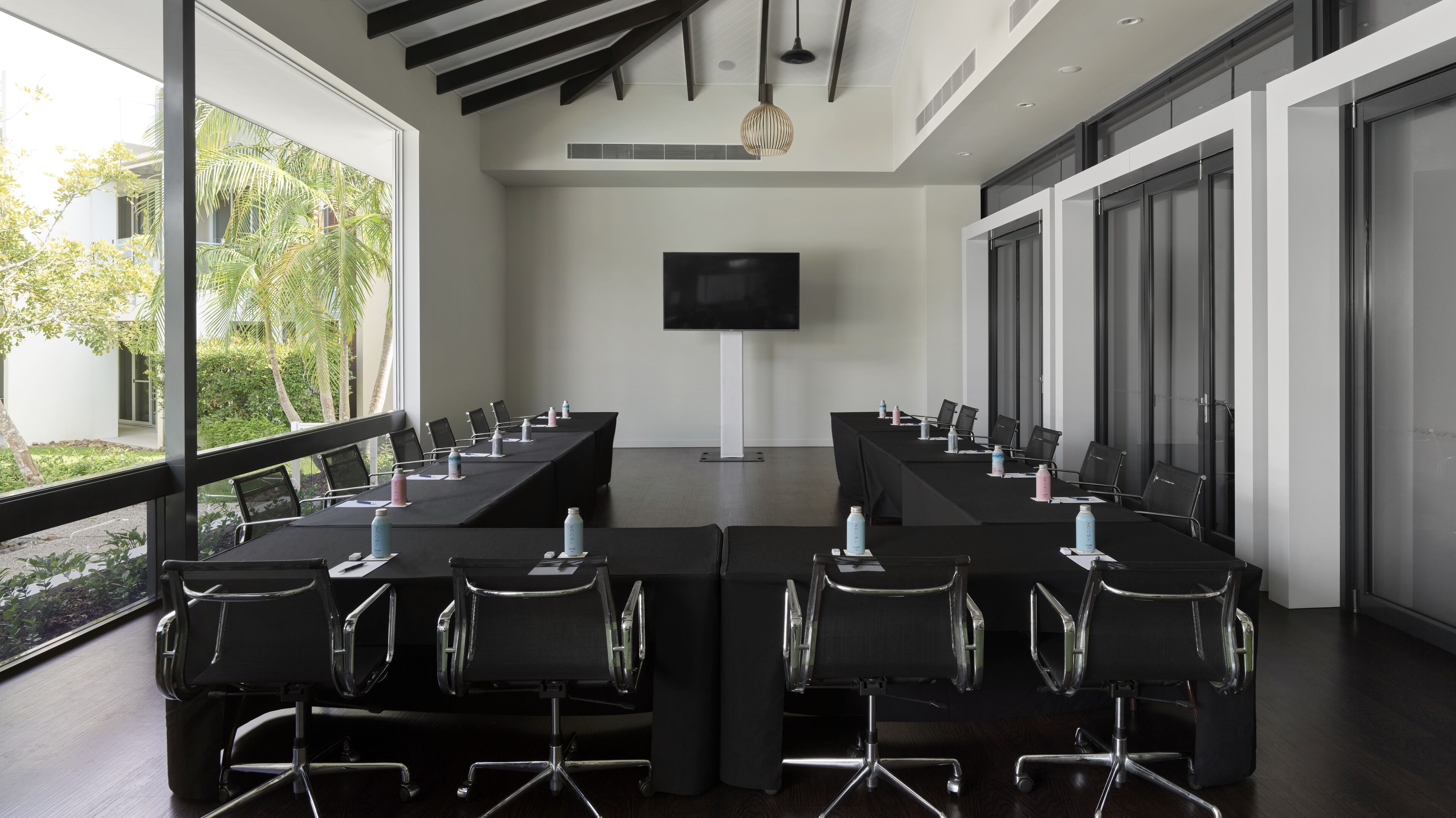 A conference room with a long table, chairs, a TV screen, and large windows with a view of greenery.
