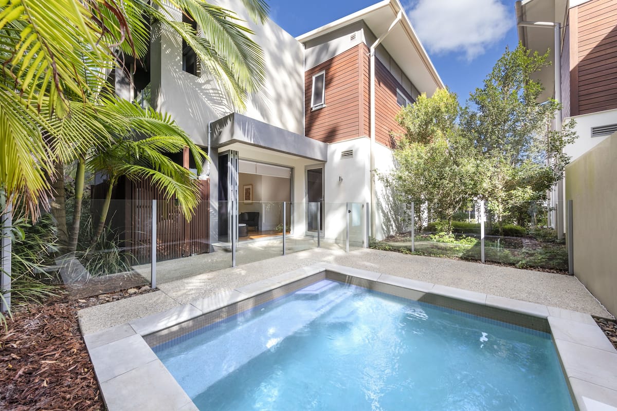 A modern residential pool area with a clear blue pool, surrounded by a wooden fence, and adjacent to a house with large windows and a balcony.