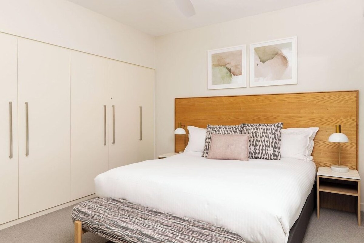 A bedroom with a large bed, a wooden headboard, two bedside tables with lamps, a bench at the foot of the bed, and a wardrobe with sliding doors.