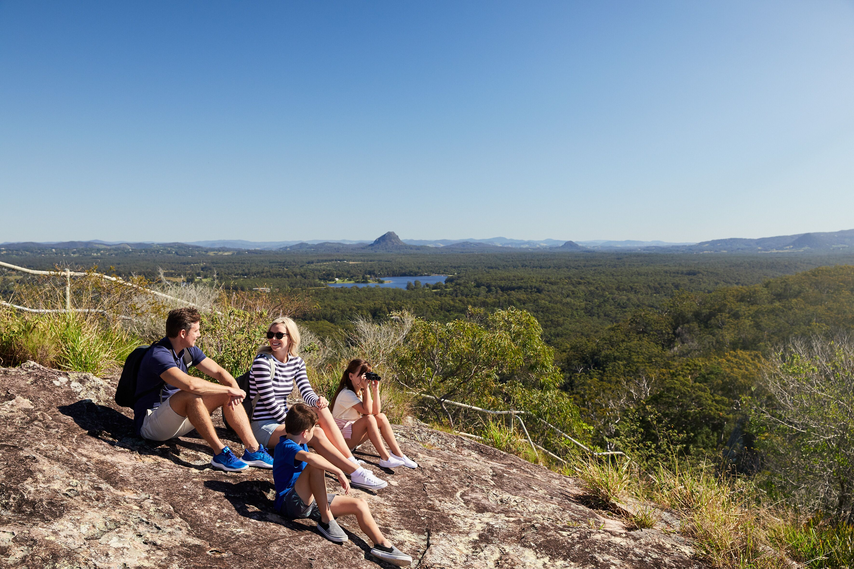 A family of four sitting on a rocky outcrop overlooking a forested valley with a distant mountain and lake.