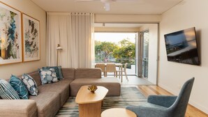 A modern living room with a sofa, coffee table, and armchair, a dining area with a table and chairs, a TV mounted on the wall, and a view through a large window. - RACV Noosa Resort (Noosa Heads)