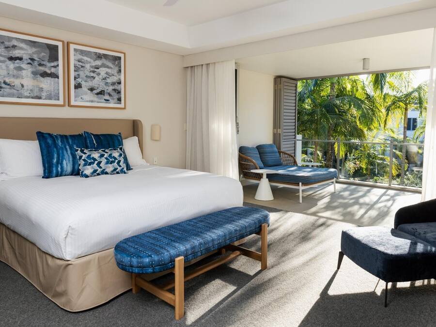 Habitación de hotel con cama, un banco, una silla y un balcón con vistas a palmeras.