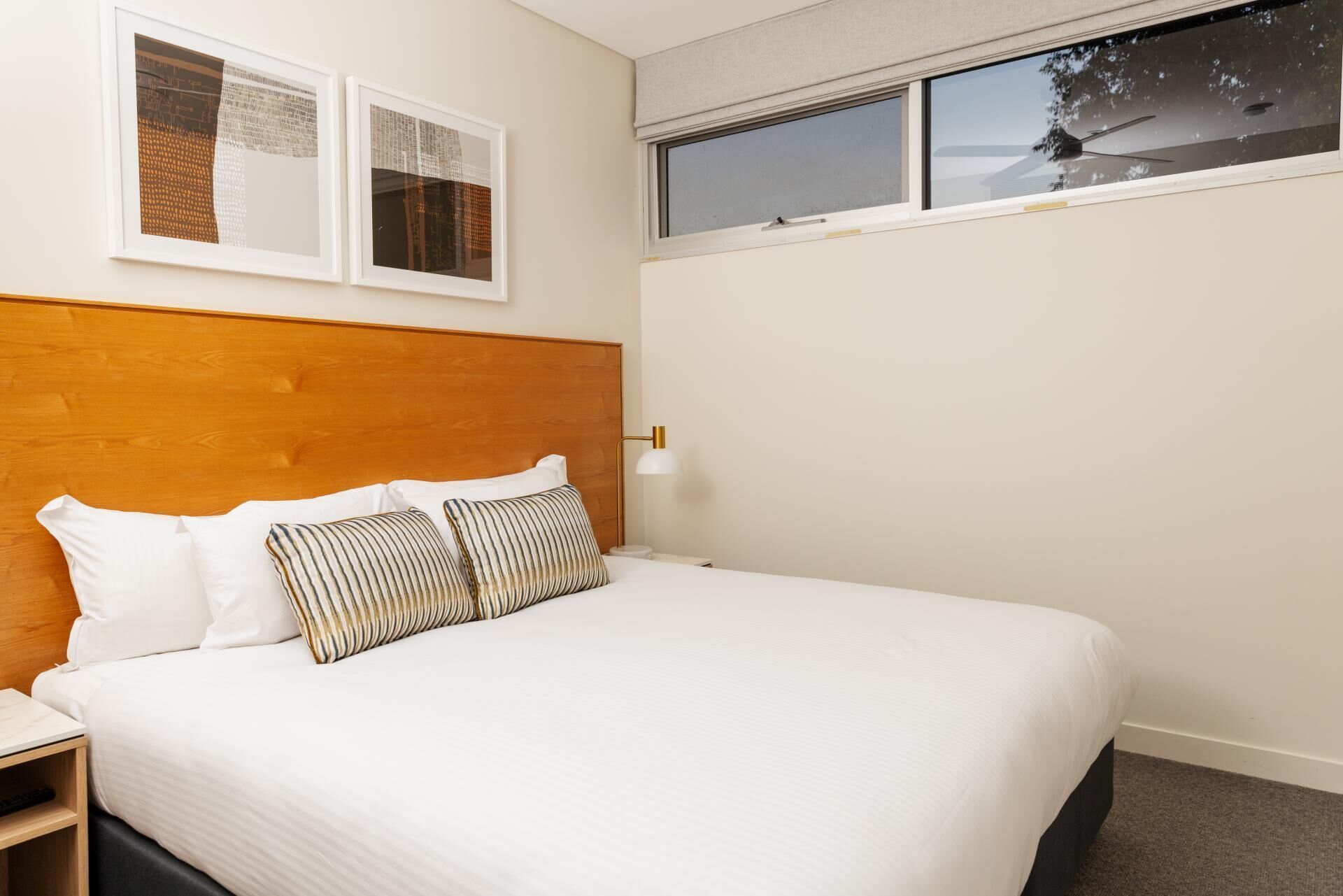 A hotel room with a wooden headboard, a bed with white bedding, striped pillows, a bedside lamp, and two framed pictures on the wall.