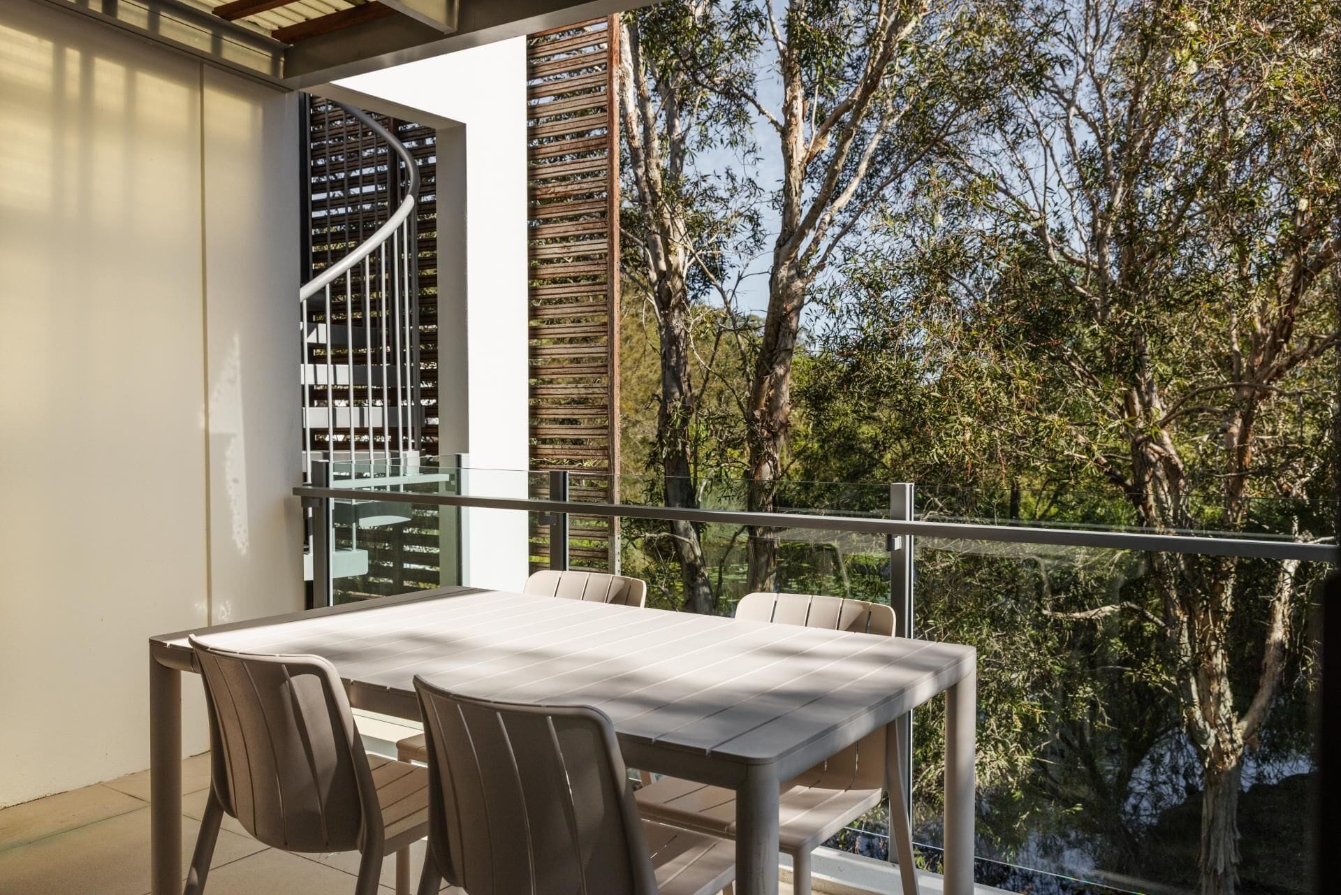 A balcony with a table and chairs, a spiral staircase, and a view of trees.