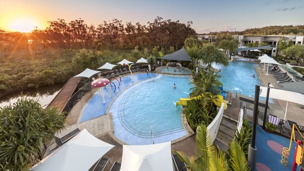 A resort pool area with a large pool, lounge chairs, umbrellas, and a view of the surrounding landscape. - RACV Noosa Resort (Noosa Heads)