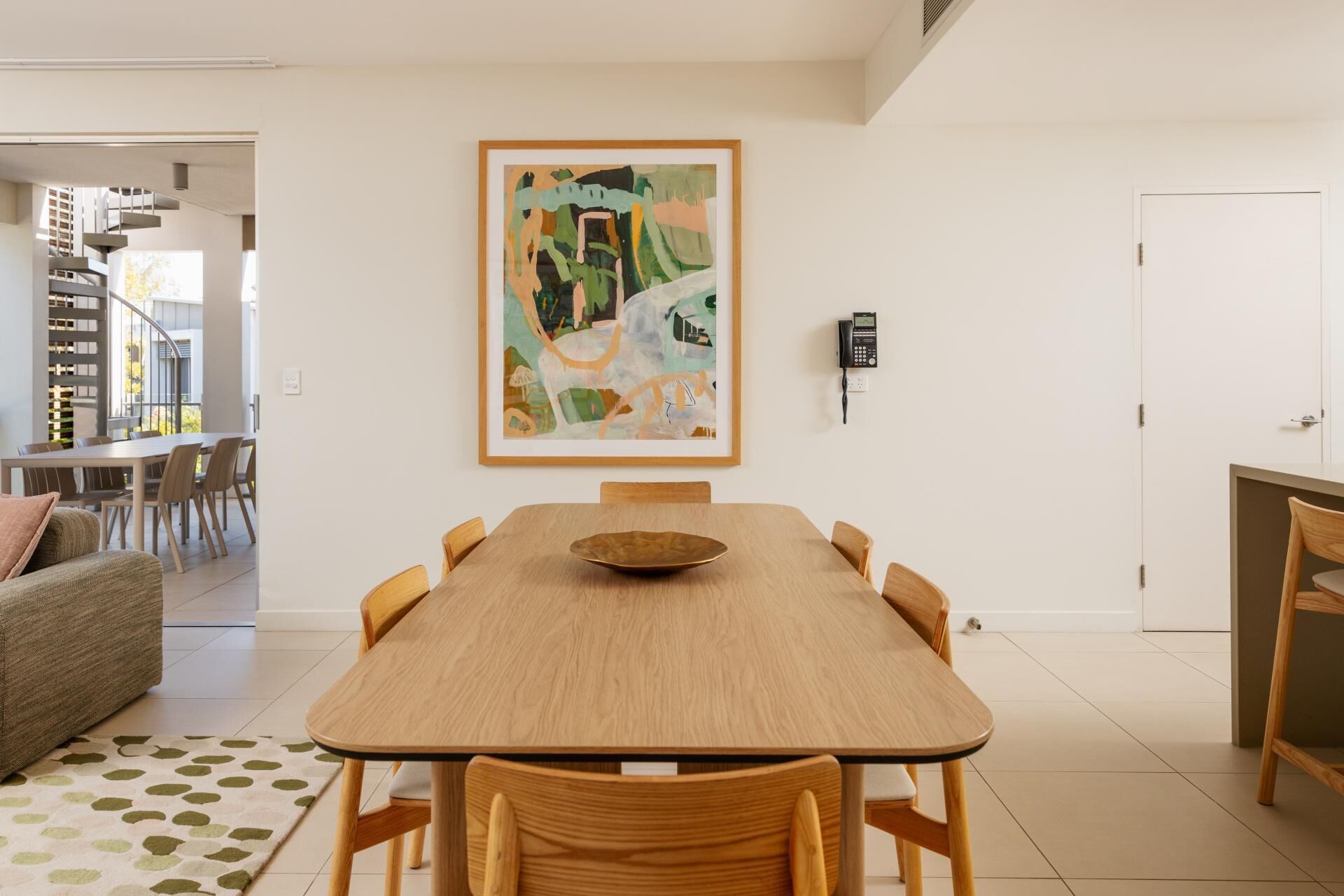 Un coin repas avec une table en bois, des chaises, un tableau abstrait encadré, un téléphone fixé au mur et un escalier en arrière-plan.