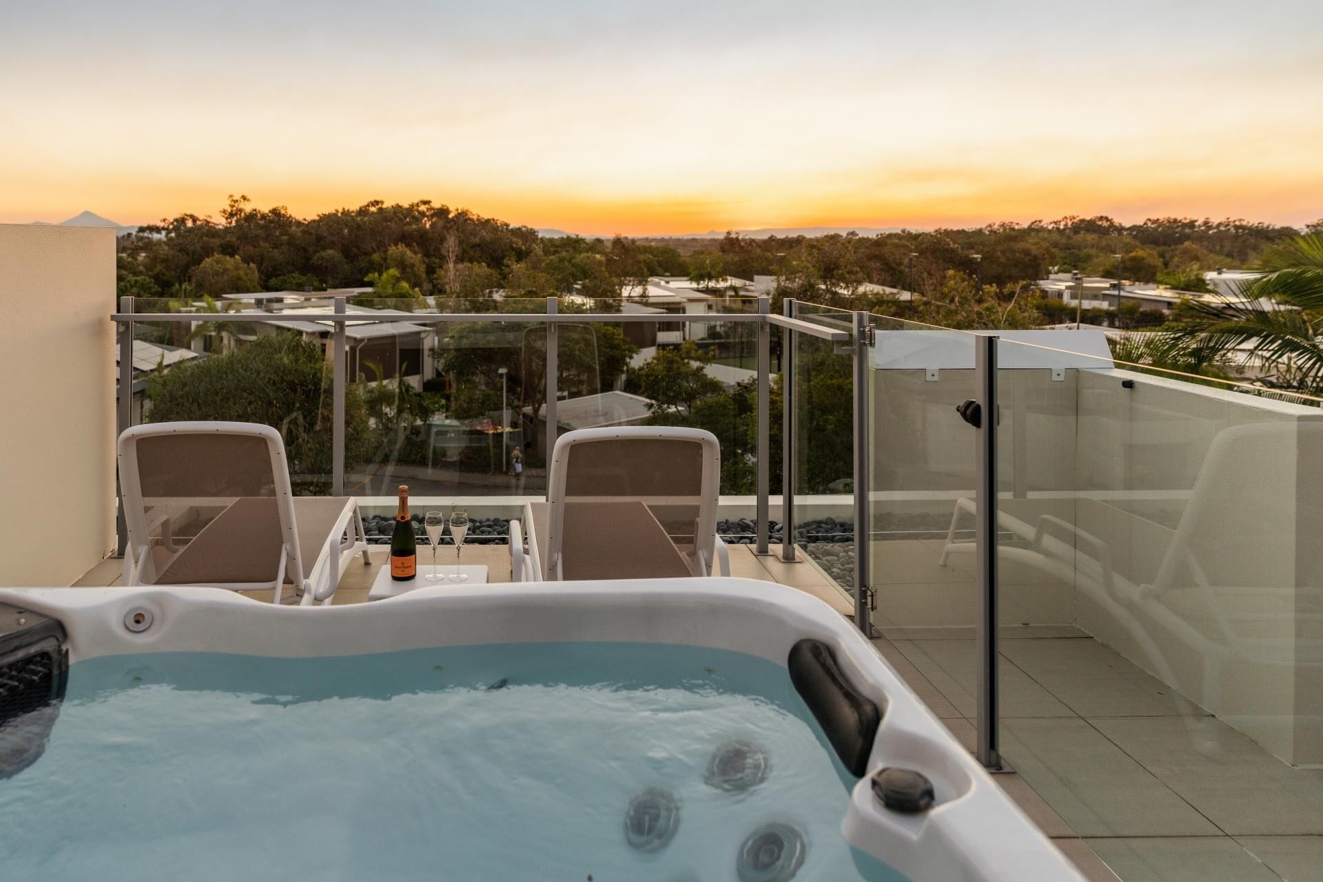 A hot tub with two chairs and a bottle of champagne, overlooking a residential area during sunset.