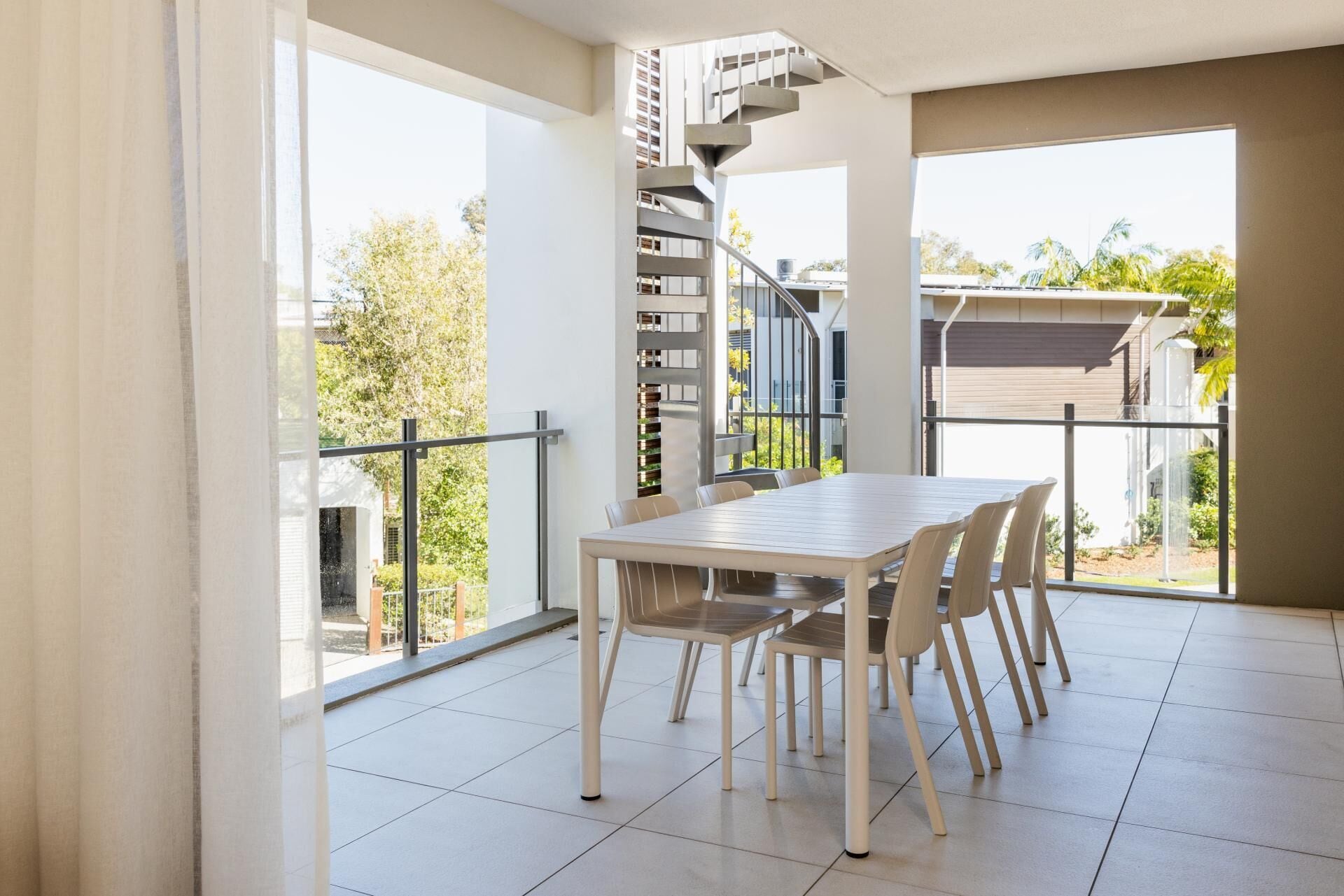 Ein modernes Interieur mit einem Esstisch und Stühlen, einer Wendeltreppe und großen Fenstern mit Blick ins Freie.