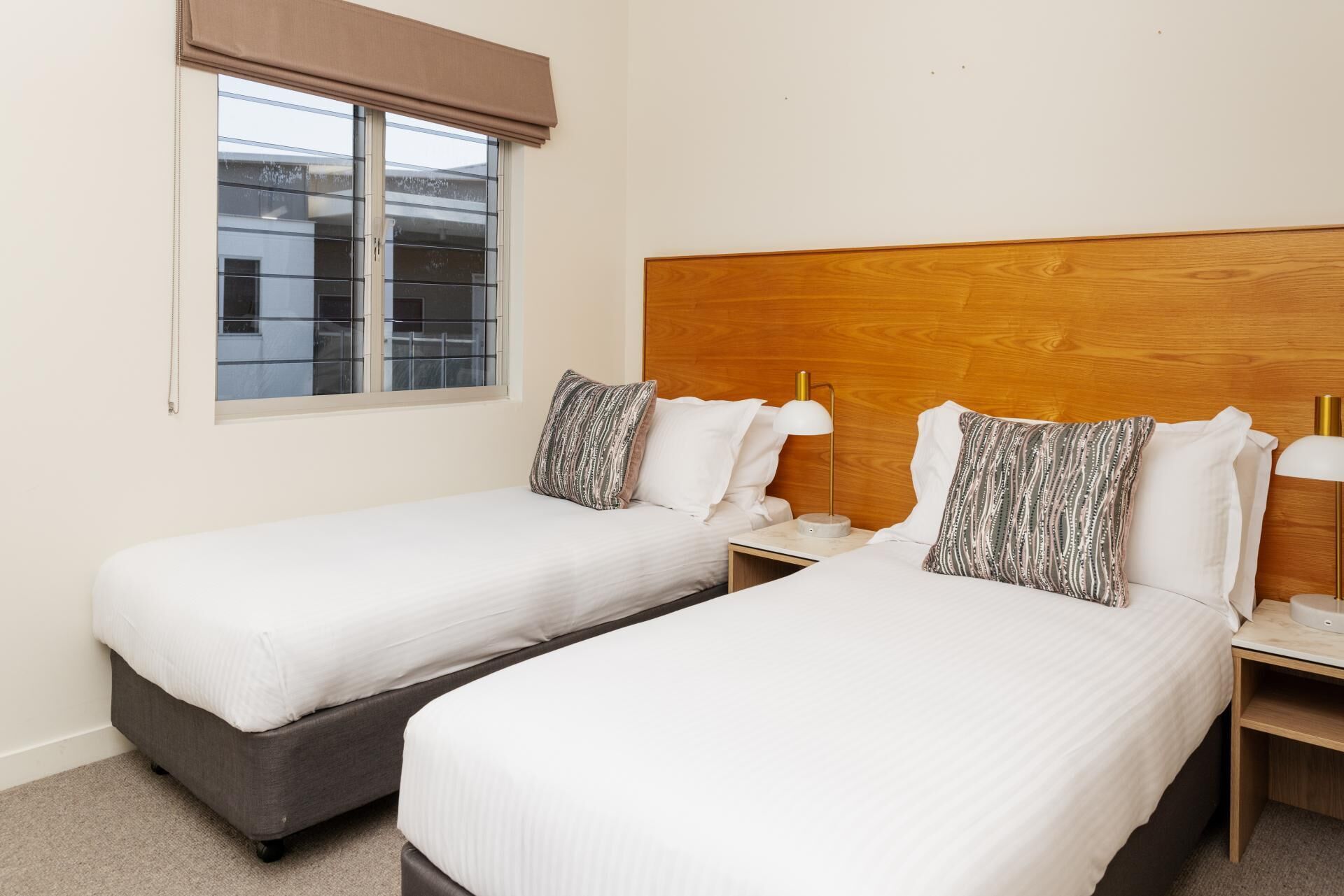 A hotel room with two single beds, a wooden headboard, a window with a blind, and a nightstand with a lamp.