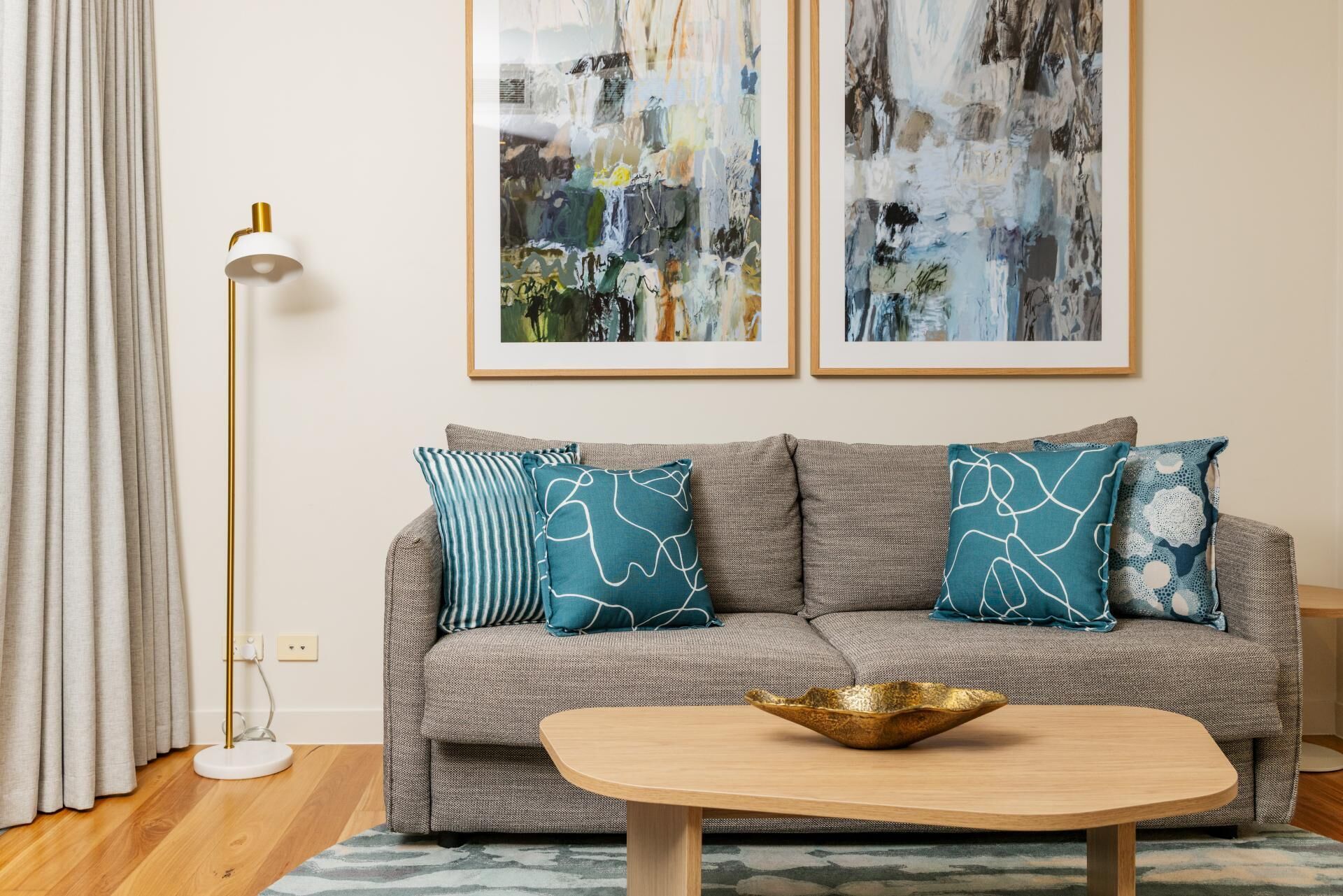 A living room with a grey sofa, teal pillows, a white floor lamp, and abstract paintings on the wall.