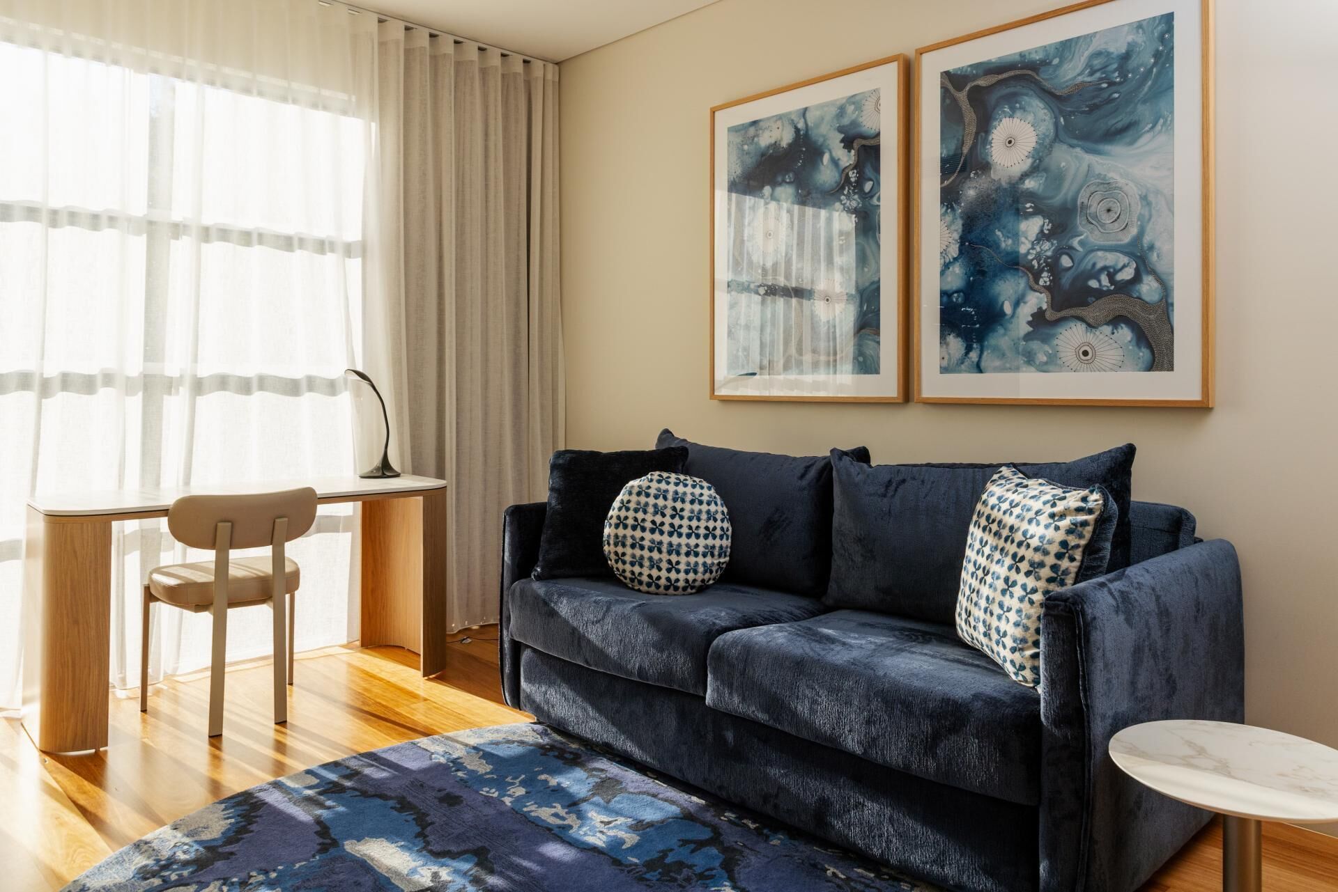 A living room with a dark blue sofa, a round coffee table, a small side table, a chair, and two framed artworks on the wall.
