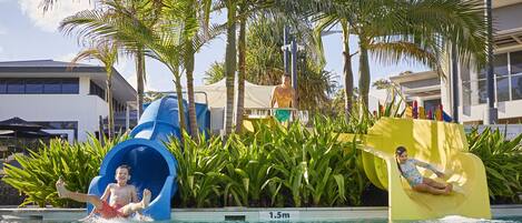 Un tobogán de agua en un área de piscina con palmeras y un edificio al fondo.
