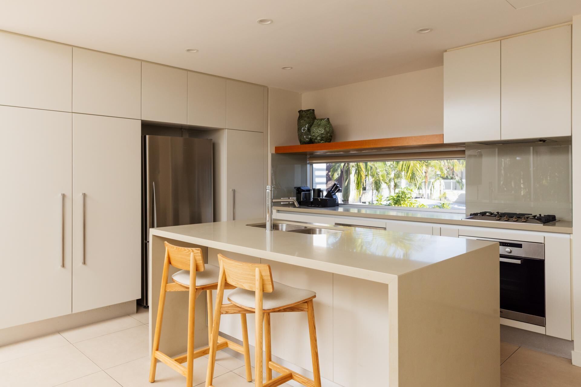 A modern kitchen with a central island, two bar stools, stainless steel appliances, and a view of palm trees outside.
