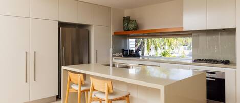 Cocina moderna con una isla central, dos taburetes de bar, electrodomésticos de acero inoxidable y vistas a palmeras.