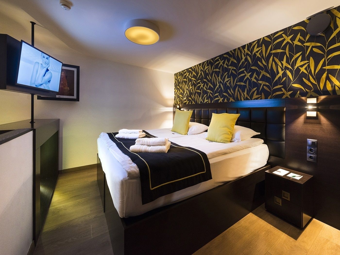 A modern hotel room with a large bed, a flat-screen TV mounted on the wall, and a decorative wall panel.