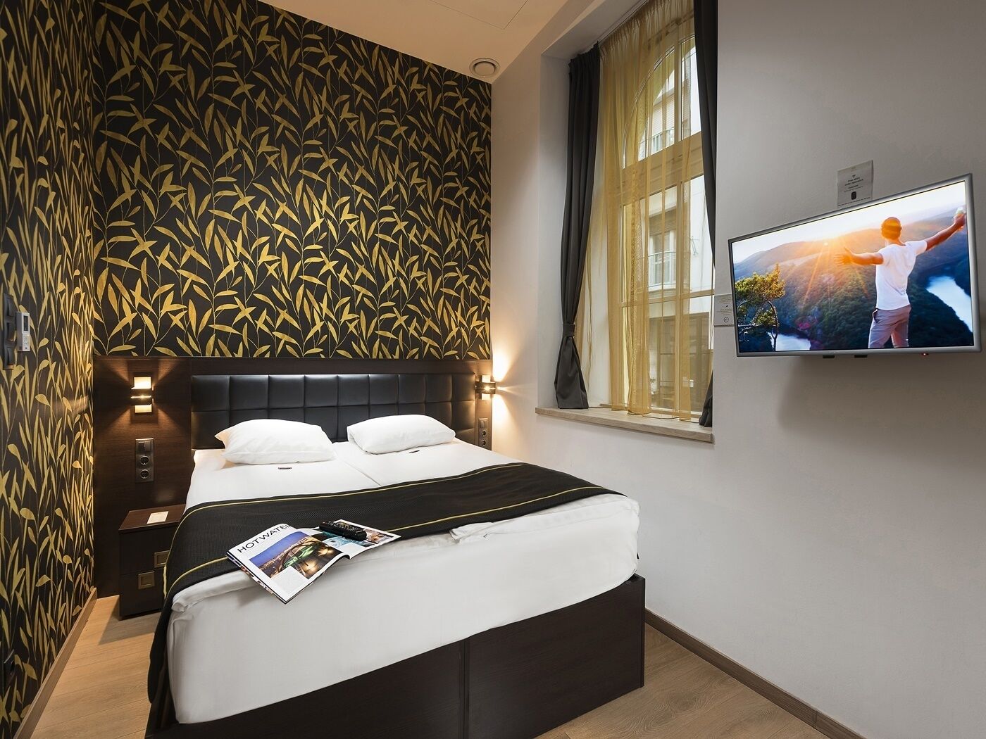 Una habitación de hotel con una cama, un televisor en la pared y un ventanal con cortinas.