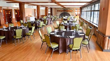 A large banquet hall with round tables and green chairs, wooden floors, and a stone fireplace.