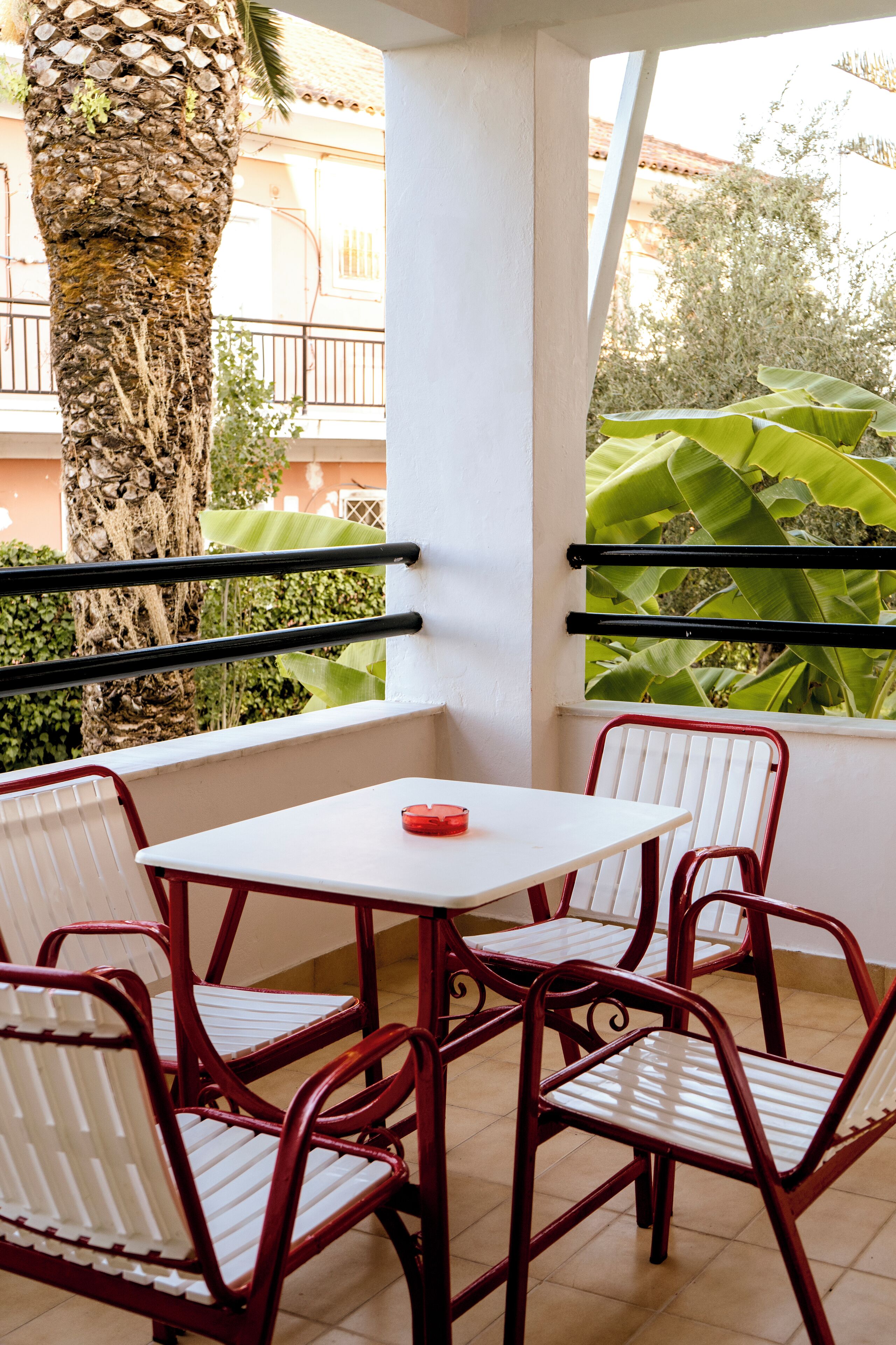 Un rincón con asientos, una mesa blanca y sillas de metal rojas, un cenicero rojo y vistas a una palmera y follaje.