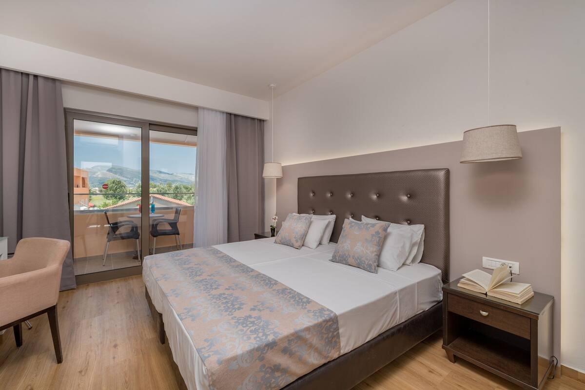 A hotel room with a large bed, a desk, and a chair. There is a balcony with a view of the mountains.