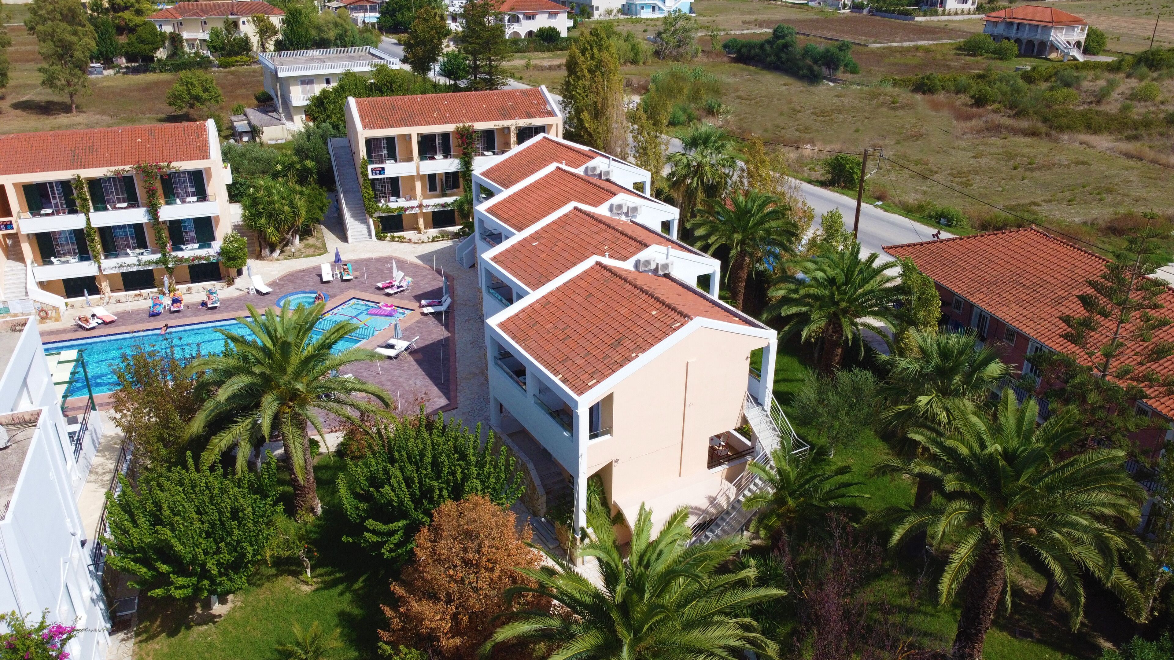 A multi-story hotel with a swimming pool, palm trees, and a red-tiled roof.