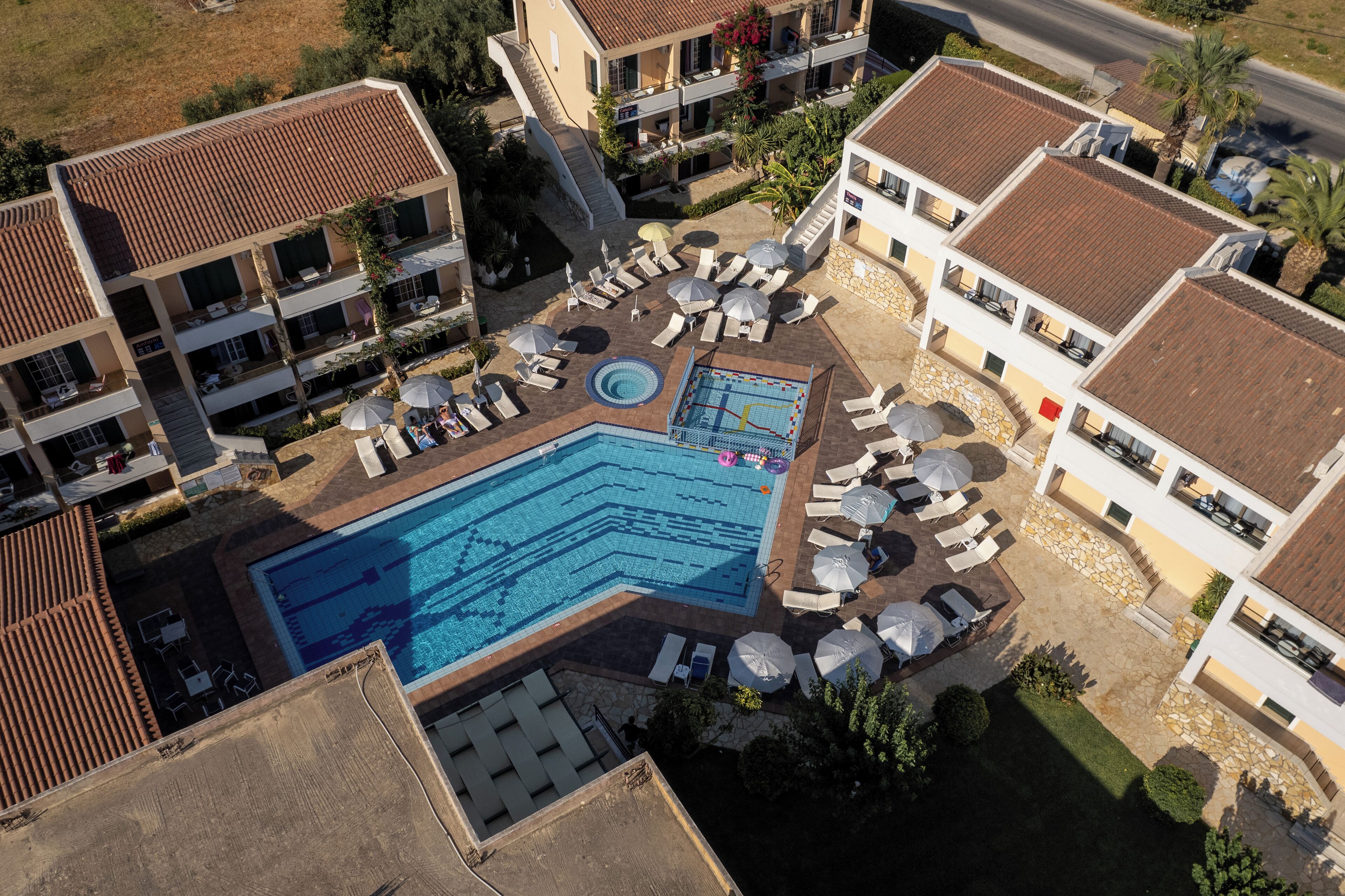Ovenfra ses et hotel med en swimmingpool omgivet af liggestole og parasoller, flere balkoner og en velplejet have.